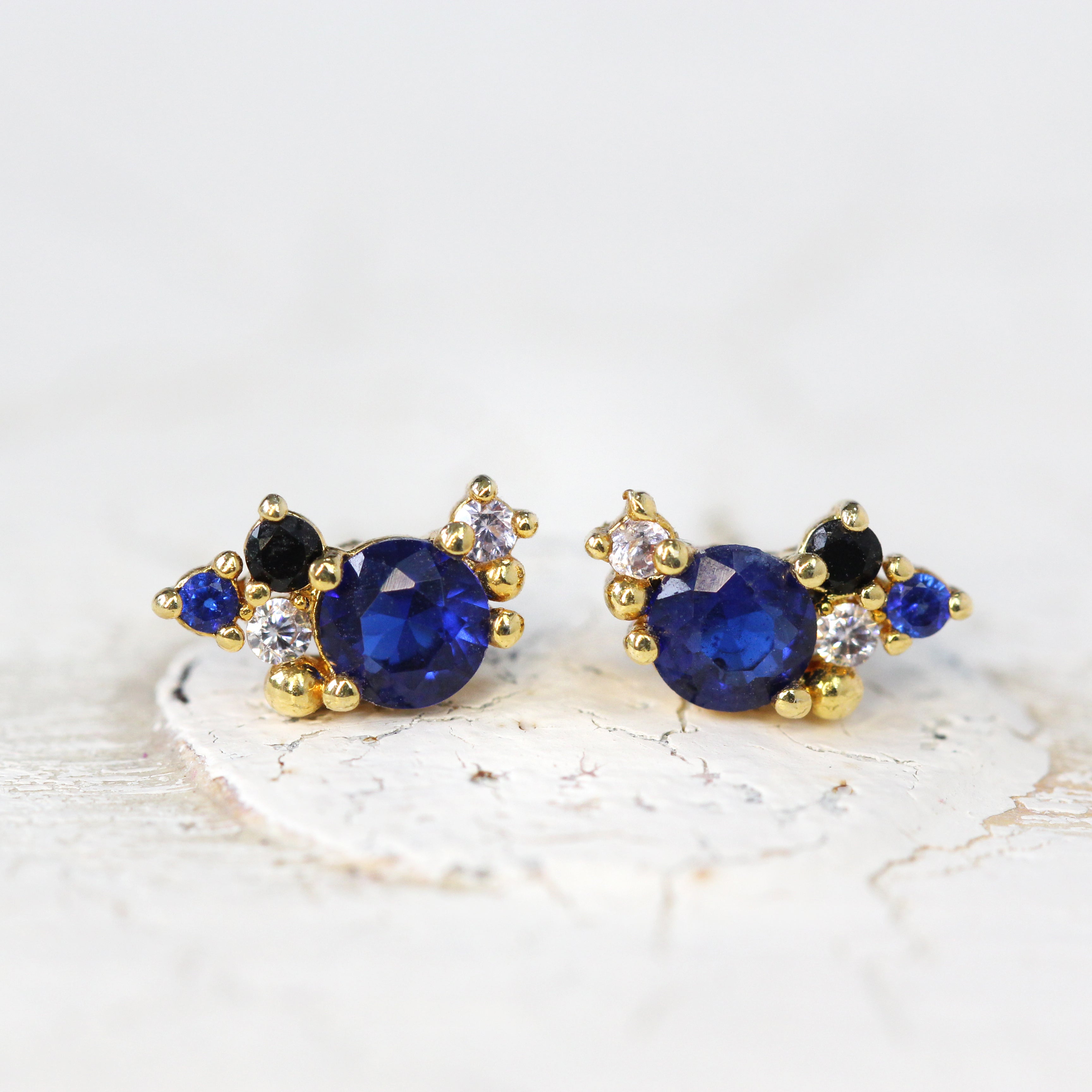 A pair of blue and clear cubic zirconia stud earrings in a cluster design featuring a main central round sparkling blue crystal stone with many smaller crystal stones set in gold surrounding it.