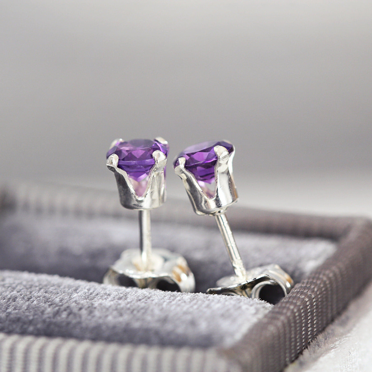 A pair of deep purple amethyst stud earrings. The amethyst gemstones are sparkling and catch the light. The stones are a brilliant round cut with lots of facets, and set into sterling silver four prong settings. Simple, minimal, round stud earrings.
