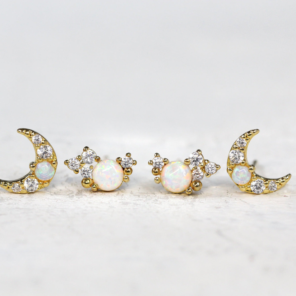 Two pairs of stud earrings side by side on a soft white background. One pair of earrings are crescent moon studs with opal and crystal stones in gold. The second pair are crystal cluster studs that resemble a galaxy constellation.