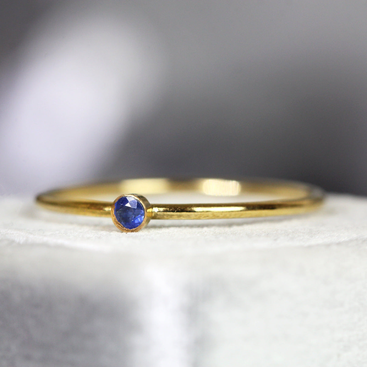Thin gold ring with a blue sapphire gemstone on white velvet with a blurred gray background