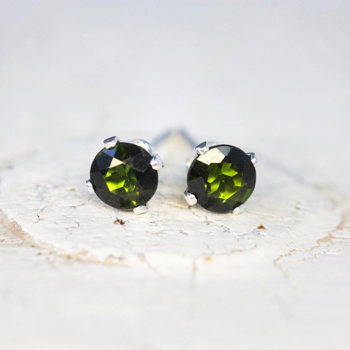 Handmade stud earrings featuring faceted dark olive green tourmaline gemstones set into a gold or silver prong setting. The earrings are round in shape, with ear post fitting and butterfly backs. The earrings are simple and minimal in style.