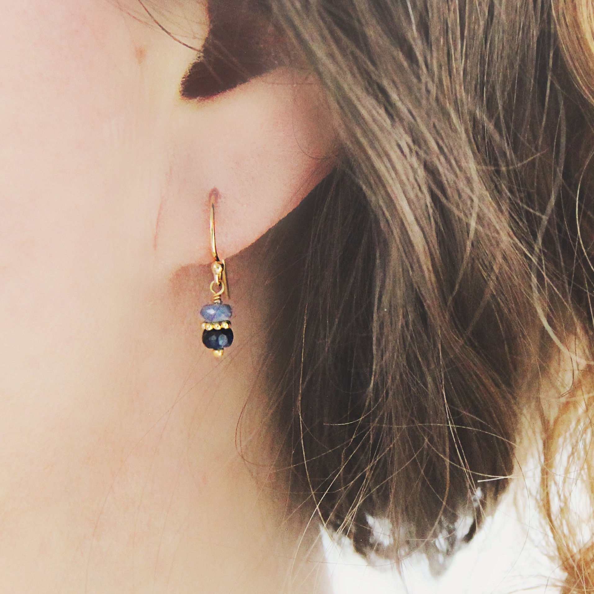 Close-up of a gold and blue sapphire drop earring worn by a person with wavy hair.