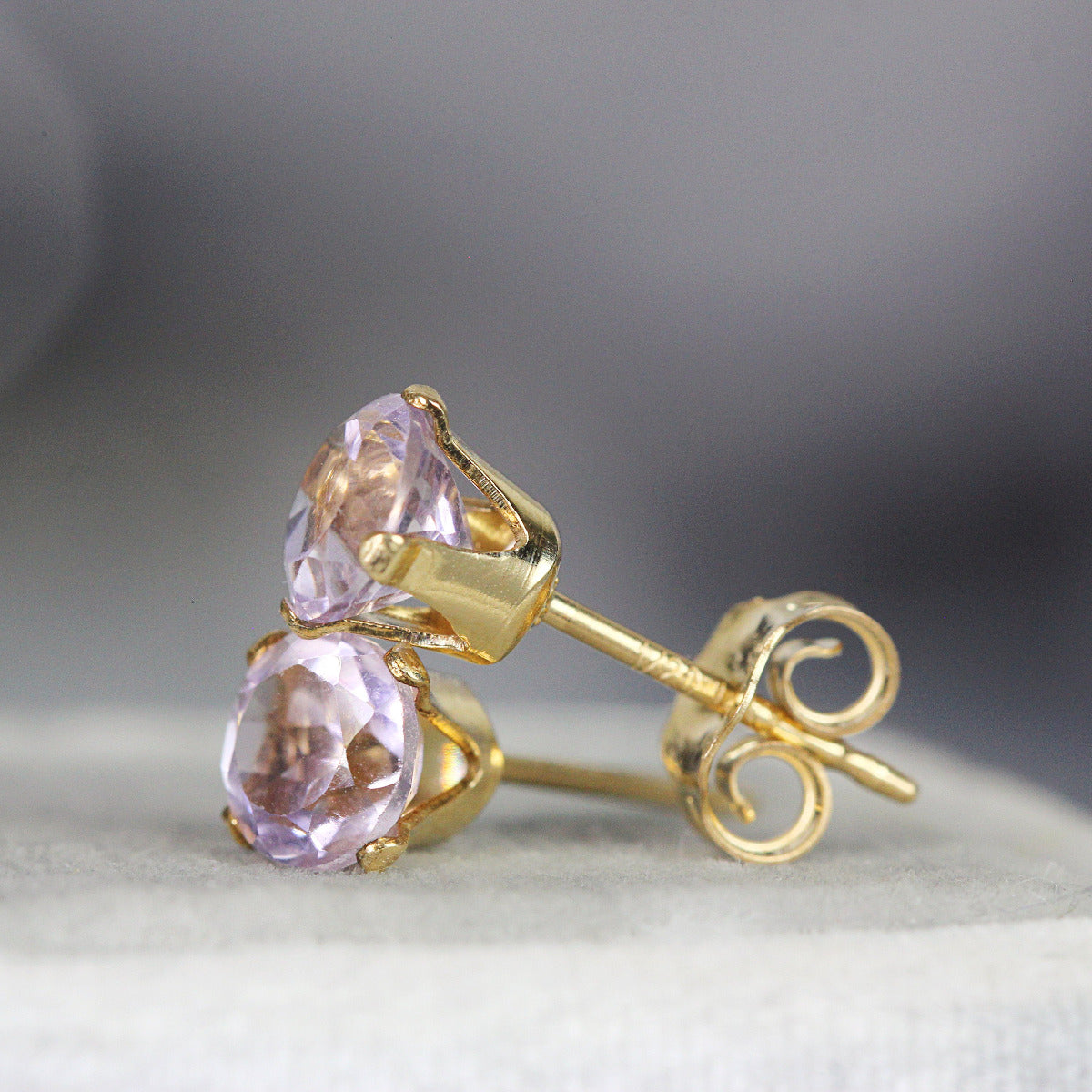 A pair of lavender purple amethyst stud earrings. The stone is round in shape with a facted brilliant round cut. The 14k gold fill ear post setting has four prongs holding the gemstone in place, and butterfly push backs to secure the stud on the ear.