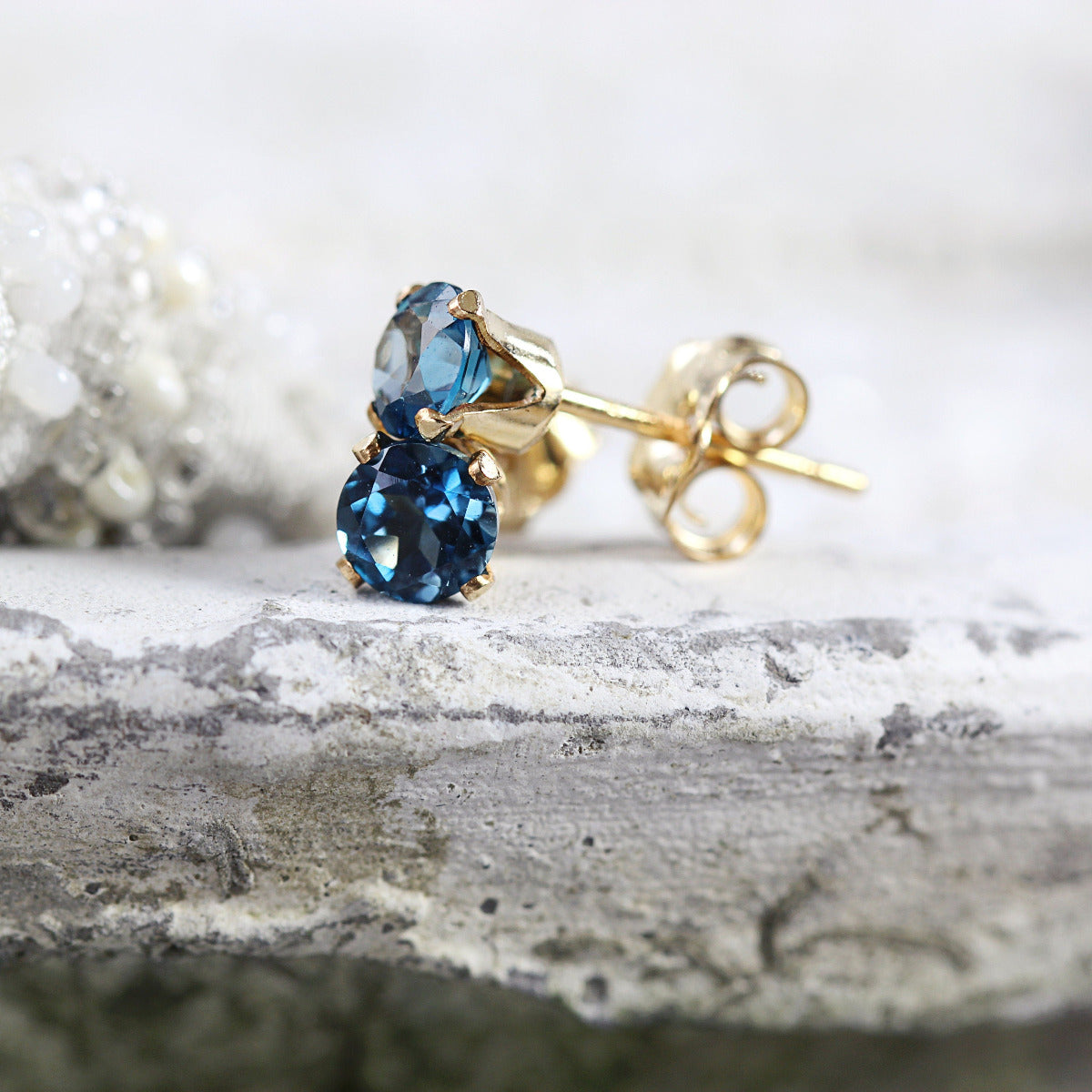 A pair of London Blue topaz stud earrings. The deep blue topaz stone is round in shape with a faceted cut. The 14k gold fill ear post setting has four prongs holding the gemstone in place, and butterfly push backs to hold the stud securely on the ear