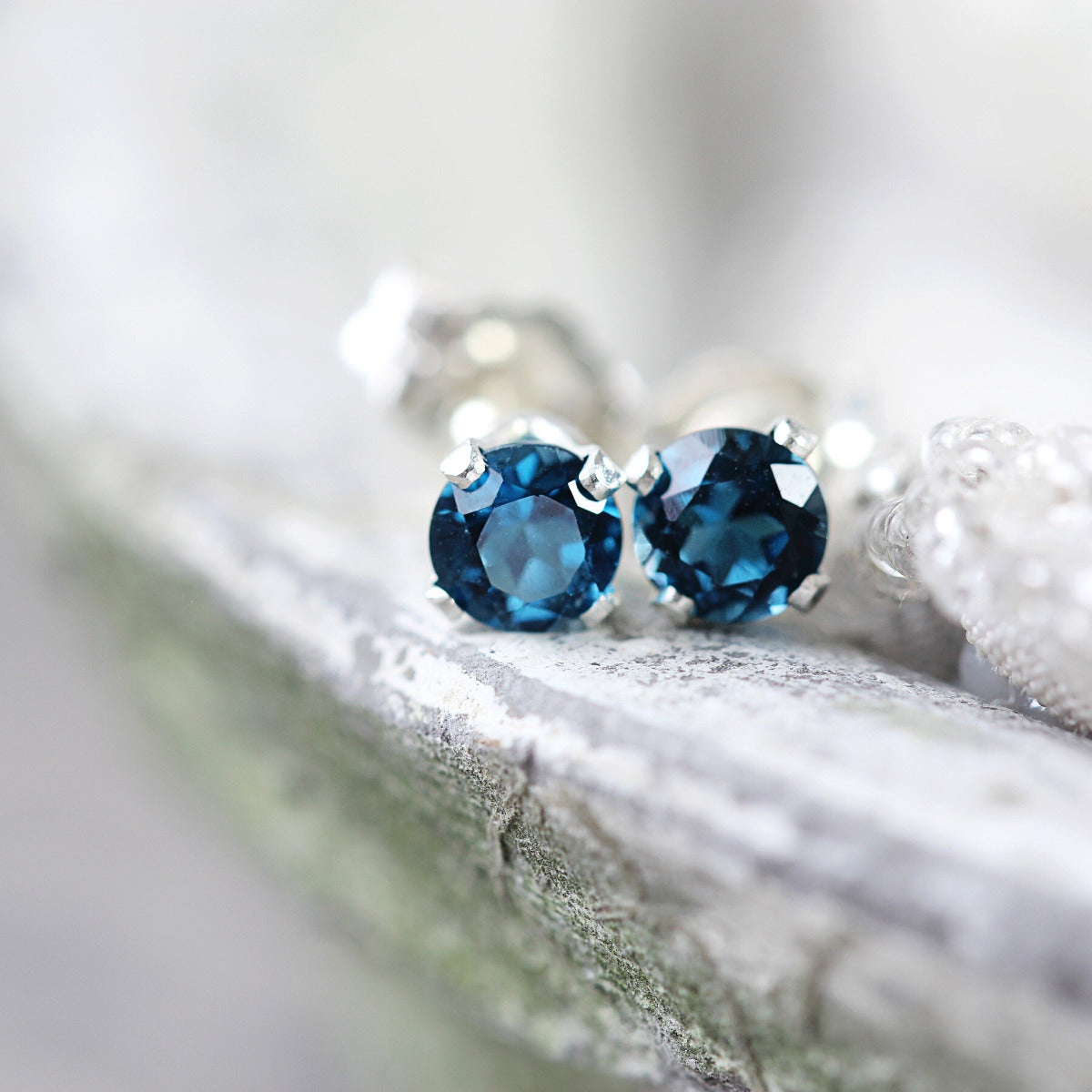 Handmade silver stud earrings featuring faceted, sparkling, brilliant round cut London Blue Topaz gemstones set into a silver prong setting. The earrings are round in shape, with ear post fitting and butterfly backs. Simple and minimal in style.