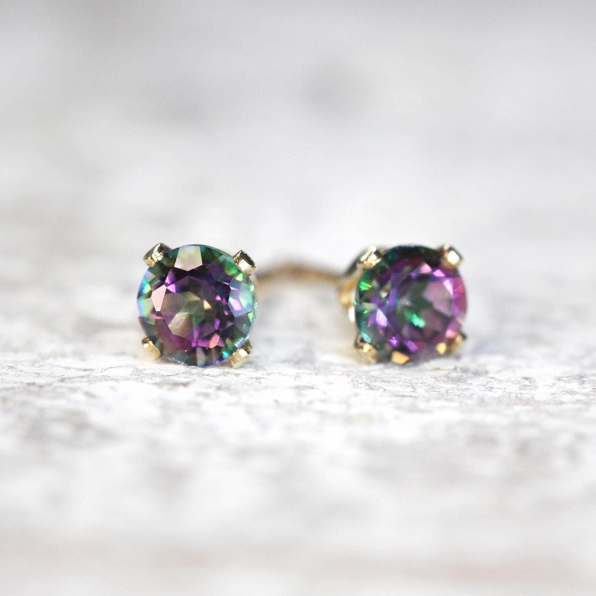A pair of stud earrings featuring colourful, vibrant kaleidoscopic mystic topaz gemstones set into a gold ear post setting. The earrings are round in shape, with ear post fitting and butterfly backs. The earrings are simple and minimal in style.