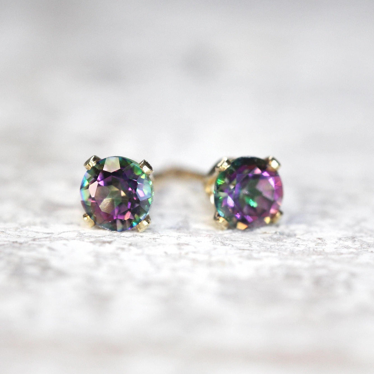 A pair of stud earrings featuring colourful, kaleidoscopic peacock shaded mystic topaz gemstones in a gold ear post setting. The earrings are round in shape, with ear post fitting and butterfly backs. The earrings are simple and minimal in style