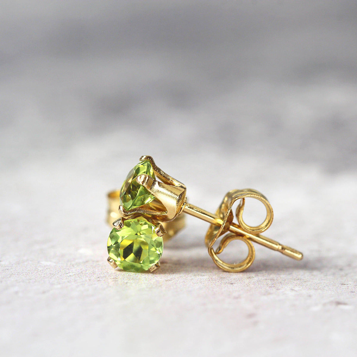 A pair of bright green peridot stud earrings. The peridot stone is round in shape with a facted cut. The 14k gold fill ear post setting has four prongs holding the gemstone in place, and butterfly push backs to hold the stud securely on the ear.