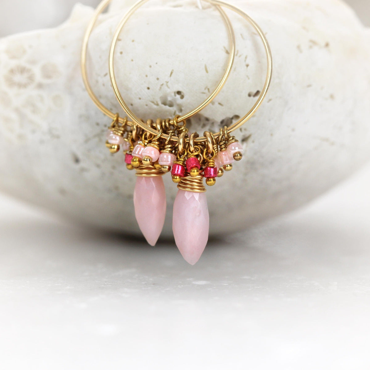 A pair of thin gold wire hoop earrings each with a long dangling pink opal teardrop and a short cluster of dangling wire wrapped matching beads in shades of baby pink and cerise pink. The style is pretty, feminine and bohemian.