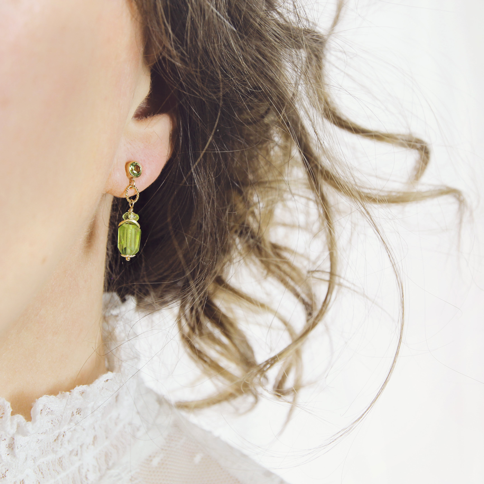 Close-up of a person wearing Raw Peridot Gemstone Drop earrings with wavy hair against a soft grey background. The handmade earrings feature raw green peridot stones that dangle from a round gemstone ear post setting. The stones are highlighted with gold accents.