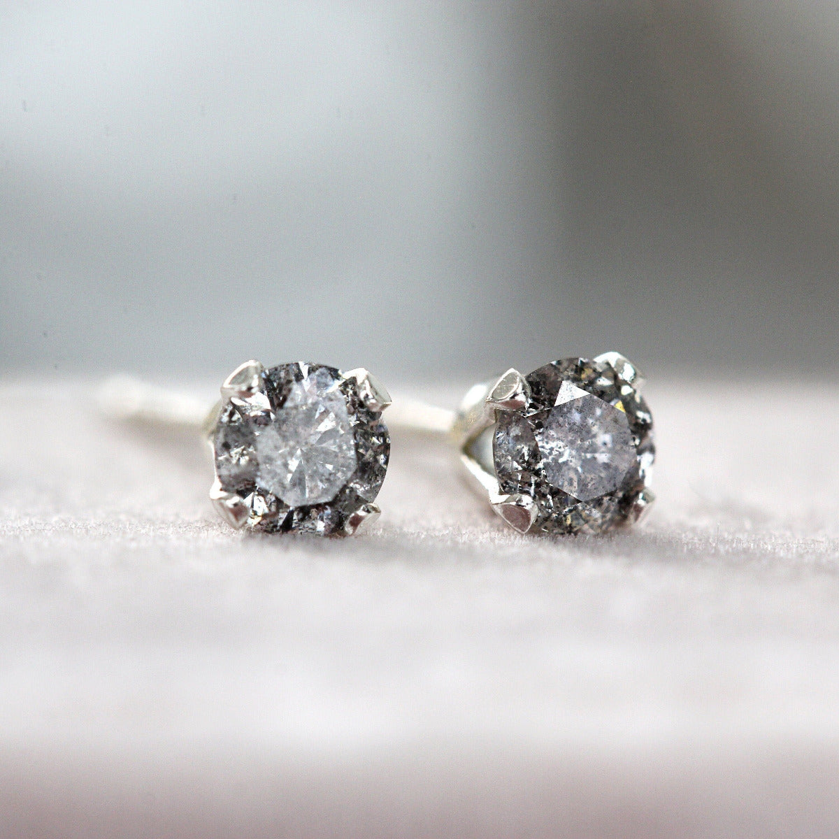 A pair of silver stud earrings featuring faceted brilliant round cut grey diamonds in a prong setting. The diamonds are called salt and pepper diamonds as they are grey with black and white flecks within that catch the light and glitter as they move.