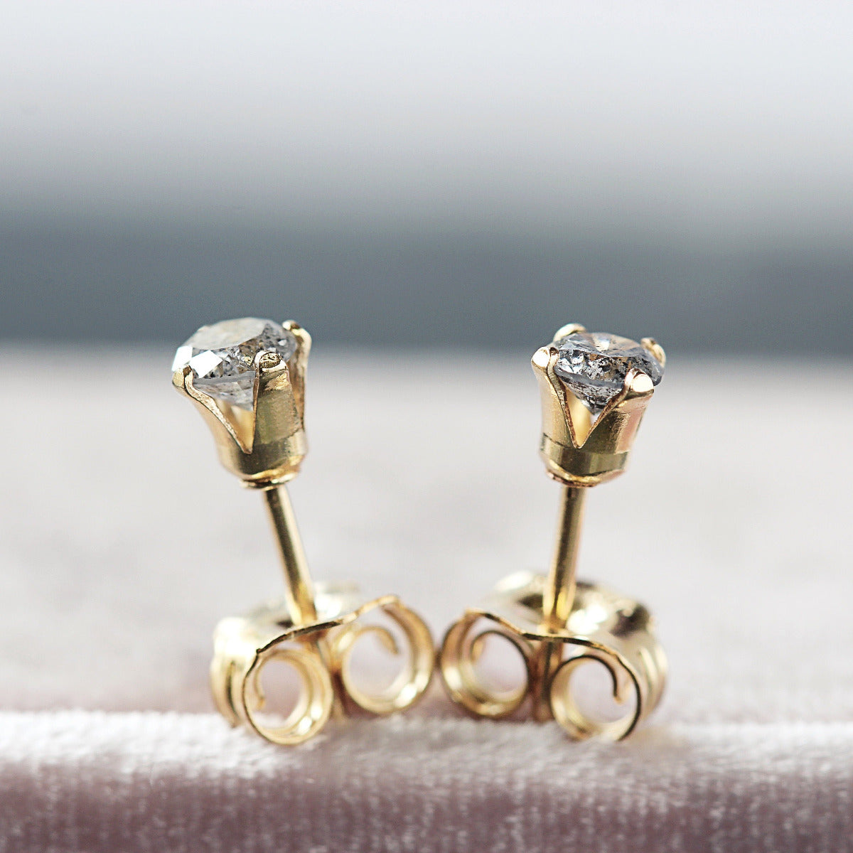 A pair of salt and pepper diamond stud earrings. The stone is round in shape with a facted brilliant round cut. The 14k gold fill ear post setting has four prongs holding the gemstone in place, and butterfly push backs to secure the stud on the ear.