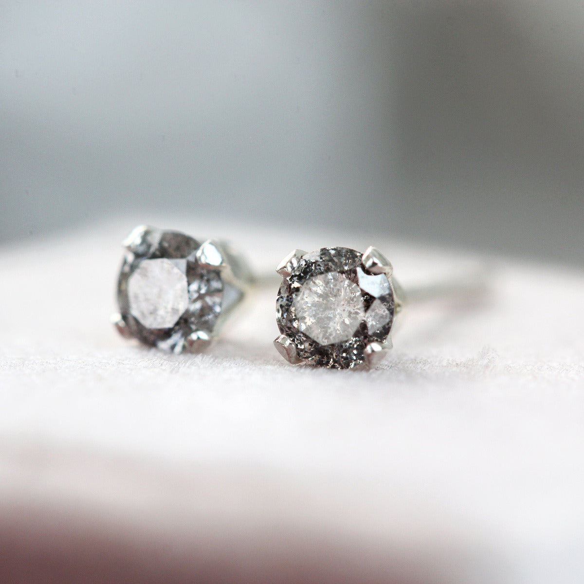A pair of grey diamond stud earrings. The diamond gemstones sparkle and glitter as they catch the light. The stones are a brilliant round cut with lots of facets, and set into sterling silver prong settings. Simple, minimal, round stud earrings.