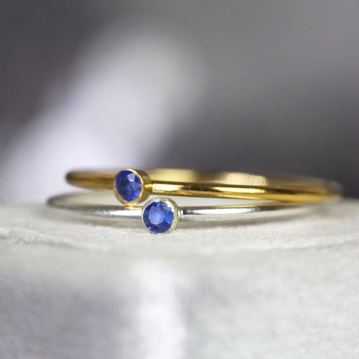 Two stacking rings with blue sapphire gemstones on a white surface. One ring is gold, the other is silver