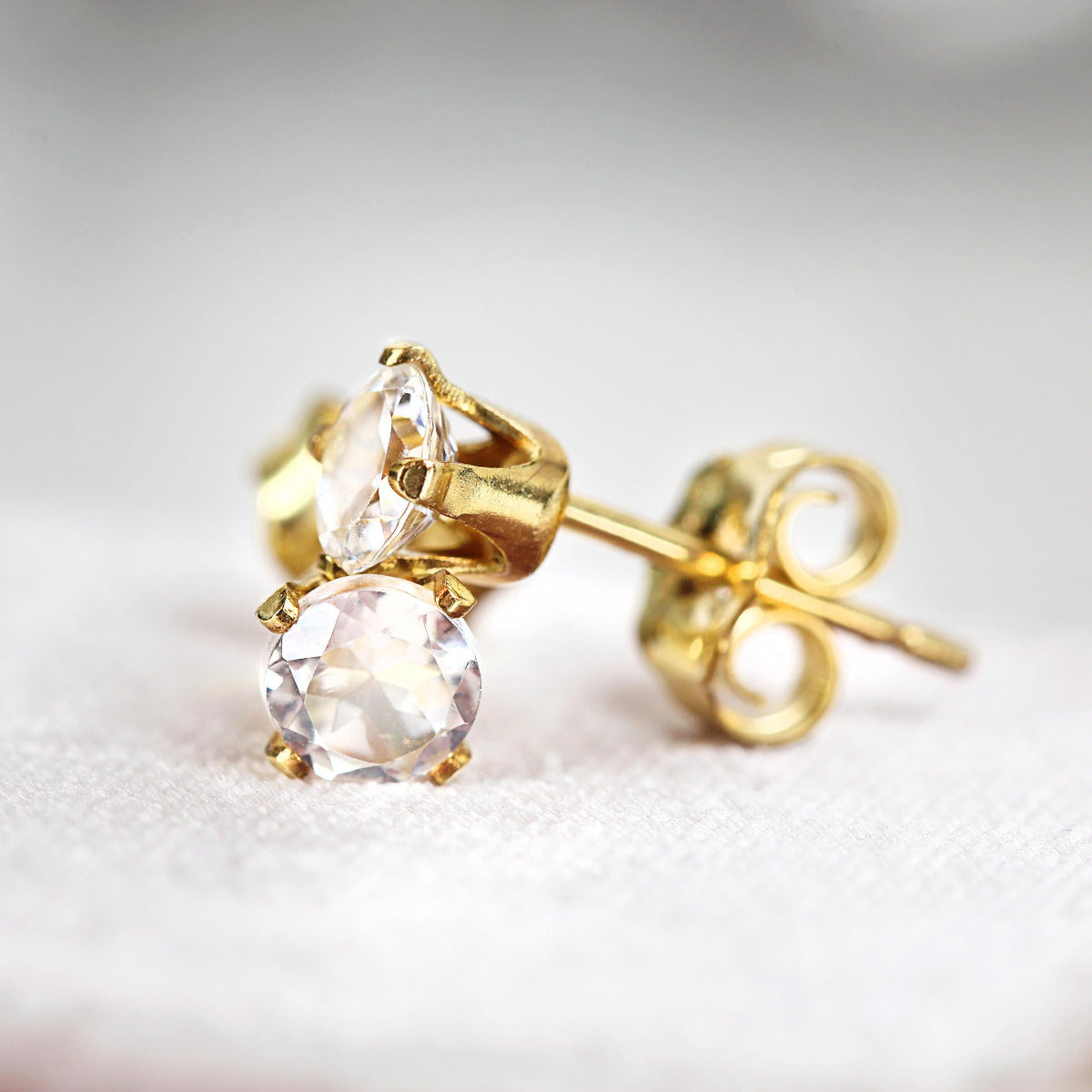 A pair of white topaz stud earrings. The topaz stones are round in shape with a facted cut. The 14k gold fill ear post setting has four prongs holding the gemstone in place, with butterfly push backs to hold the stud securely on the ear.
