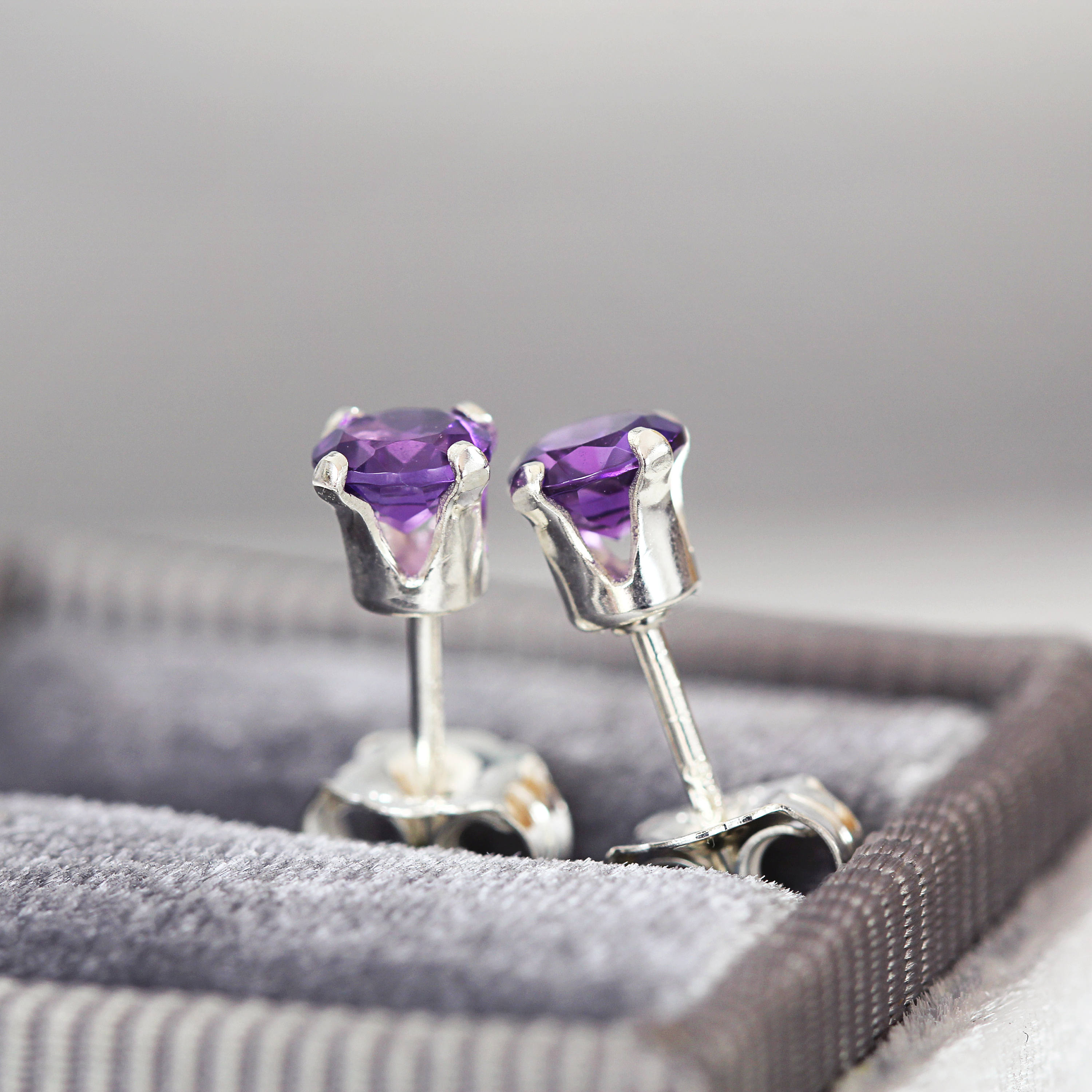A pair of dark purple amethyst stud earrings. The amethyst gemstones are sparkling and catch the light. The stones are a brilliant round cut with lots of facets, and set into sterling silver prong settings. Simple, minimal, round stud earrings.