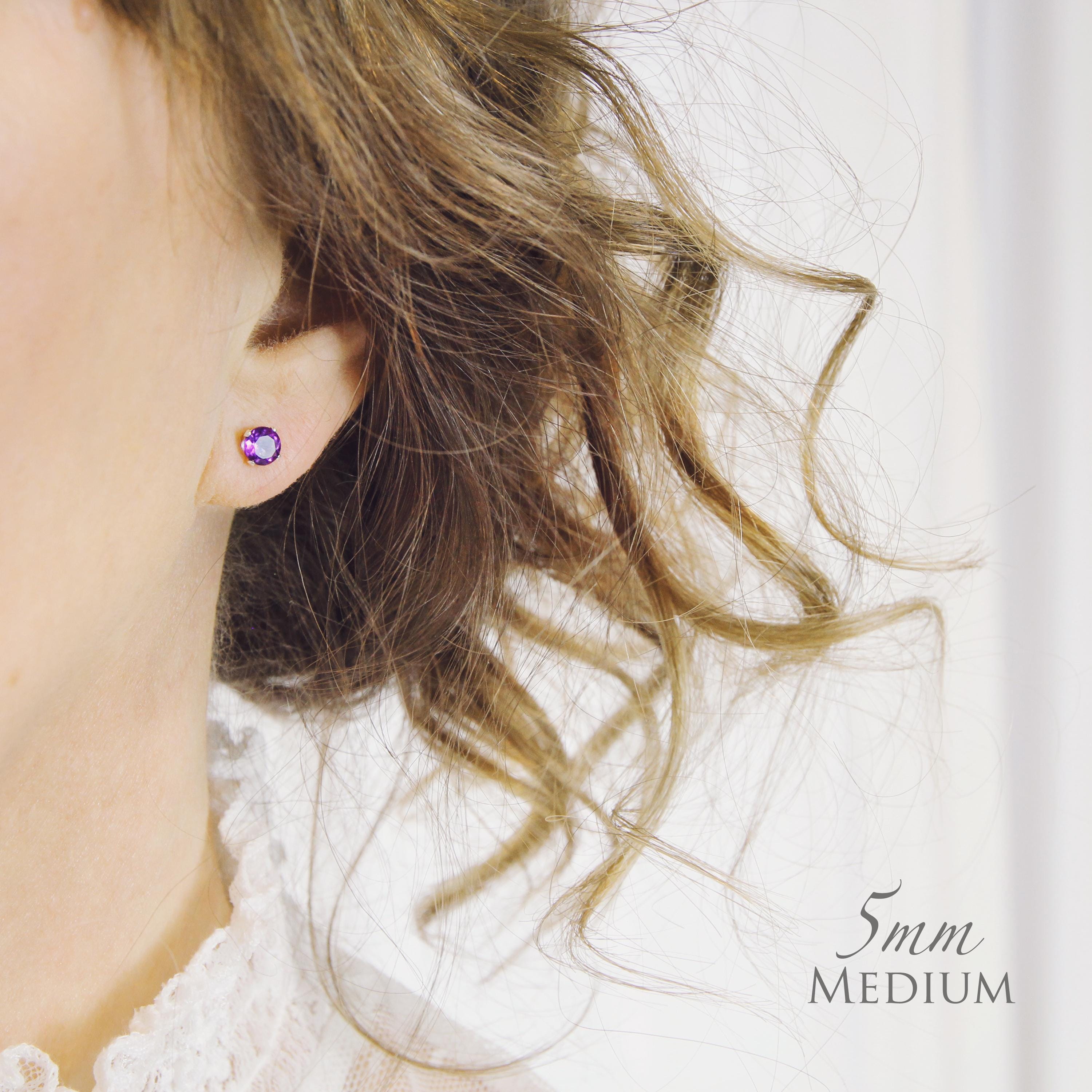 A model wears amethyst stud earrings that are small, round and dainty. The amethyst gemstone is deep purple with a faceted cut that catches the light and sparkles. The stone is set into silver ear posts. The earrings are available in gold or silver.