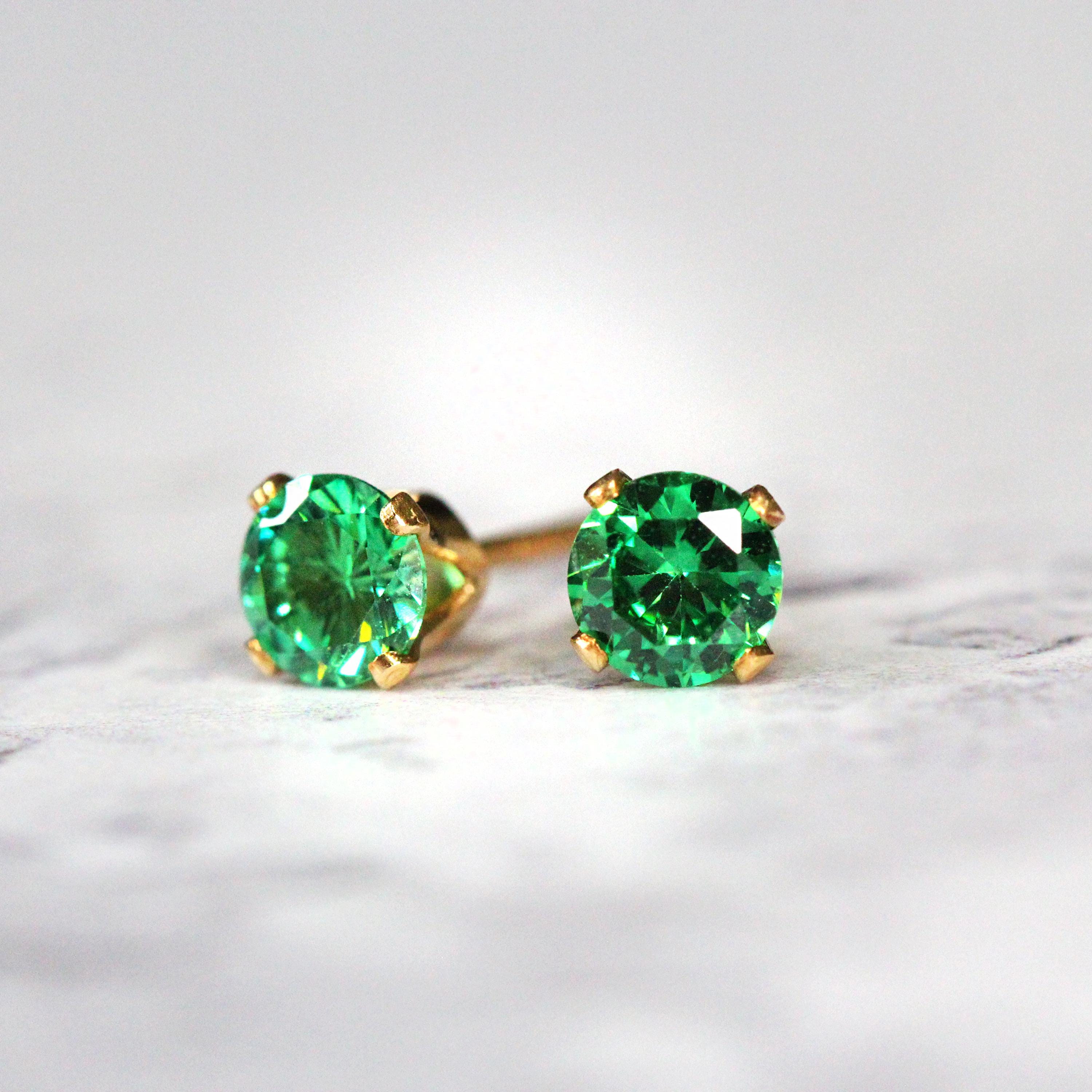 A pair of deep teal green cubic zirconia stud earrings. The cz stone is round in shape with a sparkling faceted cut. The 14k gold fill ear post setting has four prongs holding the gemstone in place, and butterfly push backs to secure the stud on the ear.