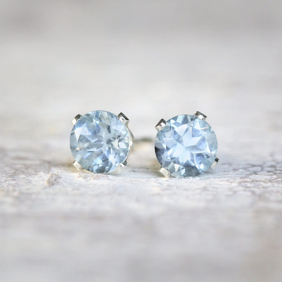 A pair of light blue aquamarine stud earrings. The aquamarine gemstones are sparkling and catch the light. The stones are round, and set into sterling silver prong settings. Simple, minimal, round stud earrings.