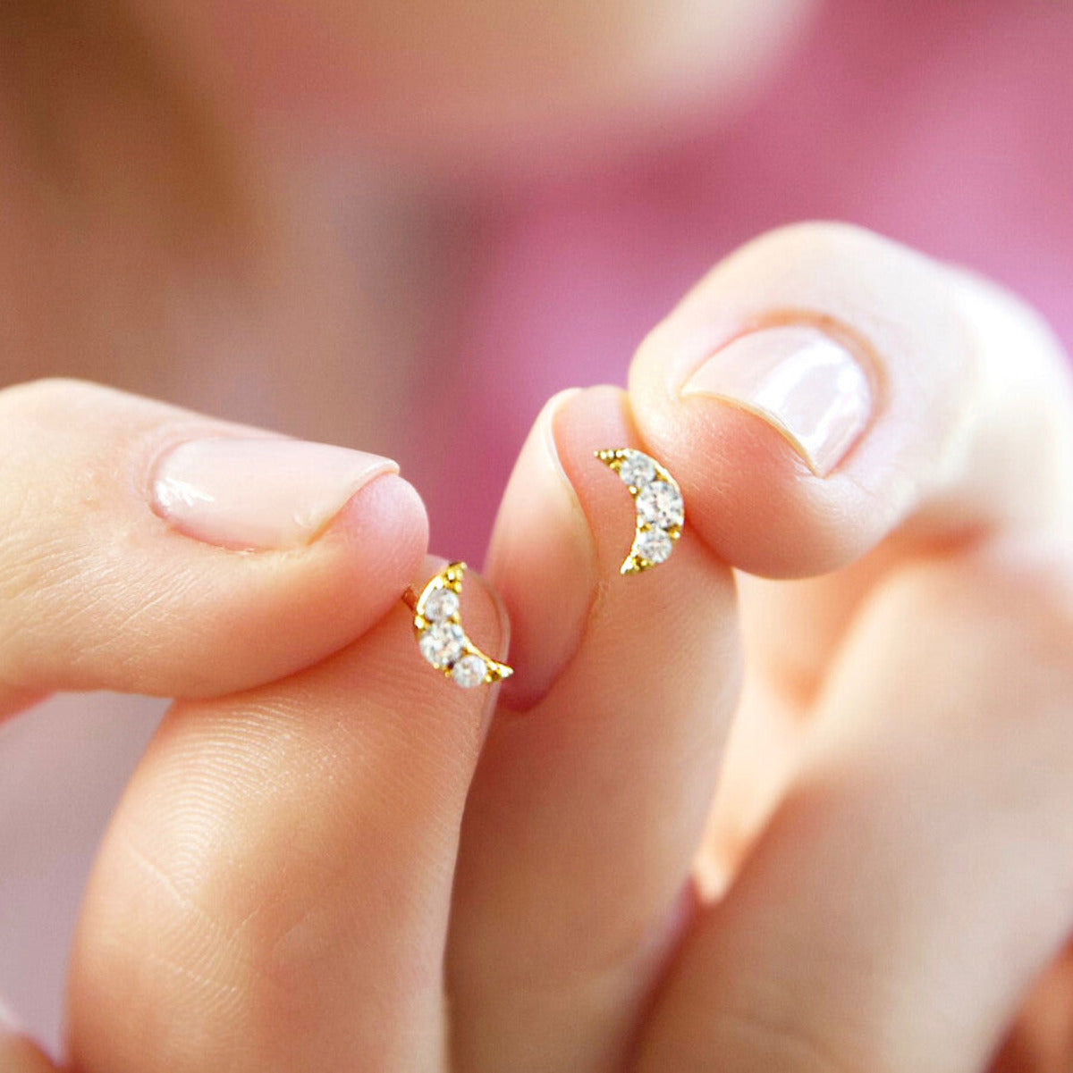 A pair of tiny gold crystal moon shaped stud earrings are held in hands of a woman