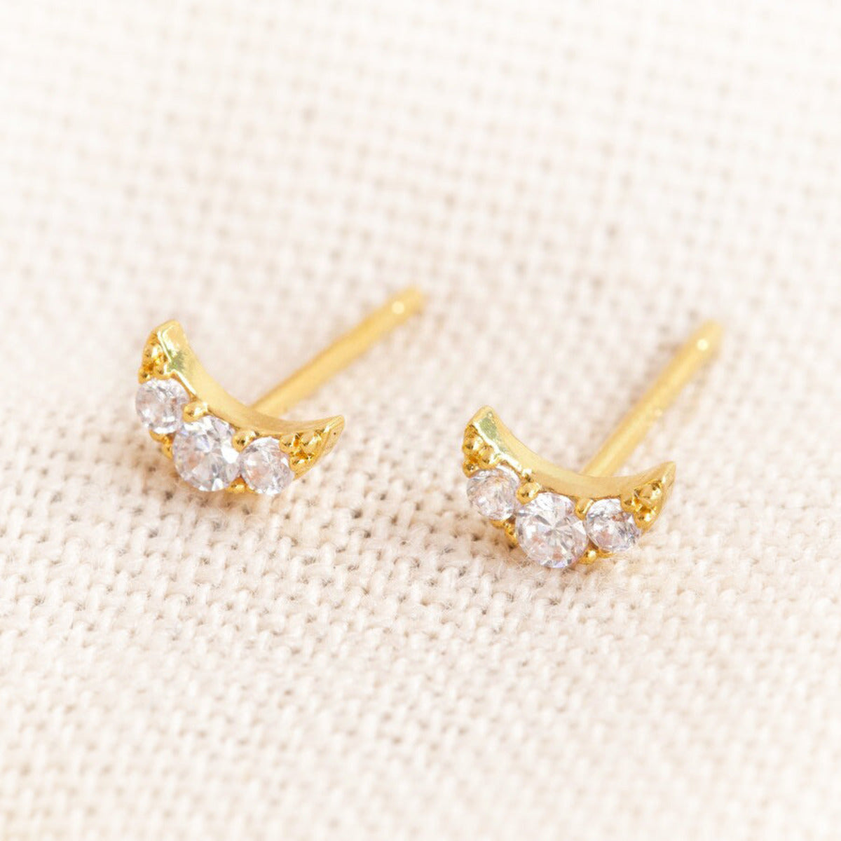 Tiny moon stud earrings with crytals set in gold against a pale fabric background