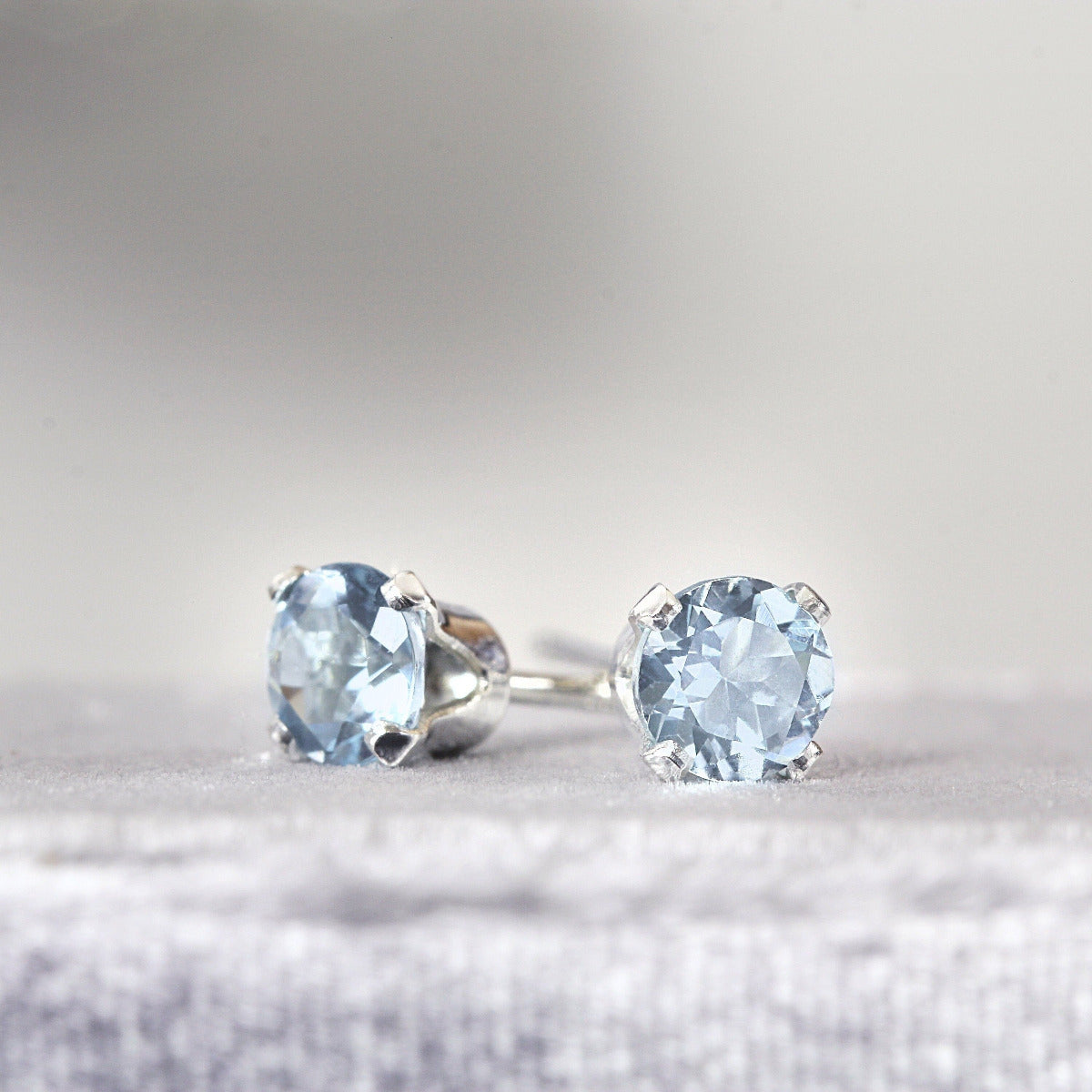 Handmade silver stud earrings featuring faceted, sparkling, brilliant round cut light blue aquamarine gemstones set into a silver prong setting. The earrings are round in shape, with ear post fitting and butterfly backs. Simple and minimal in style.