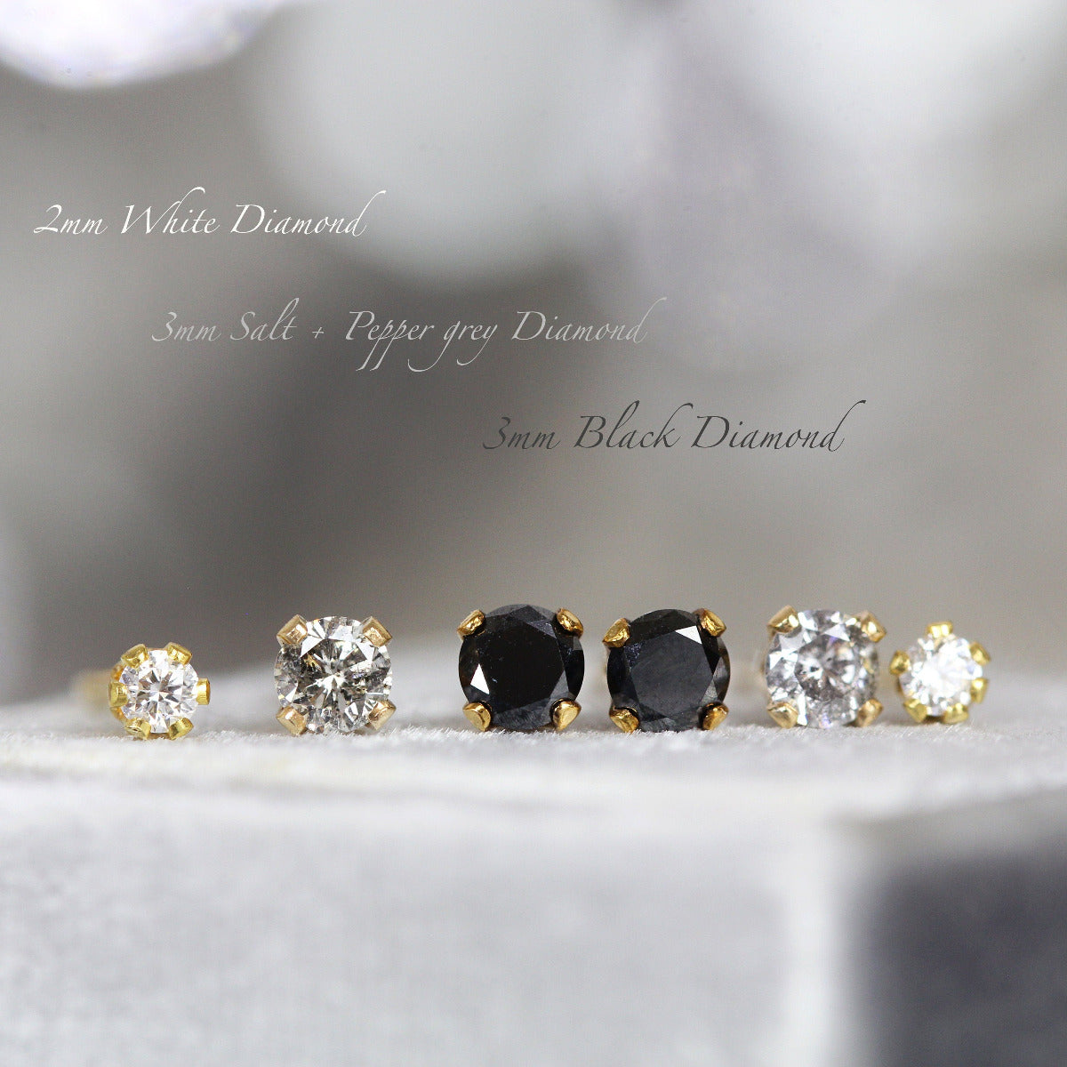 Three pairs of diamond stud earrings sit side by side. One pair of tiny 2mm white diamond studs next to one pair of 3mm grey salt and pepper diamonds next to one pair of 3mm black diamonds. The diamonds are prong set into gold ear post settings.