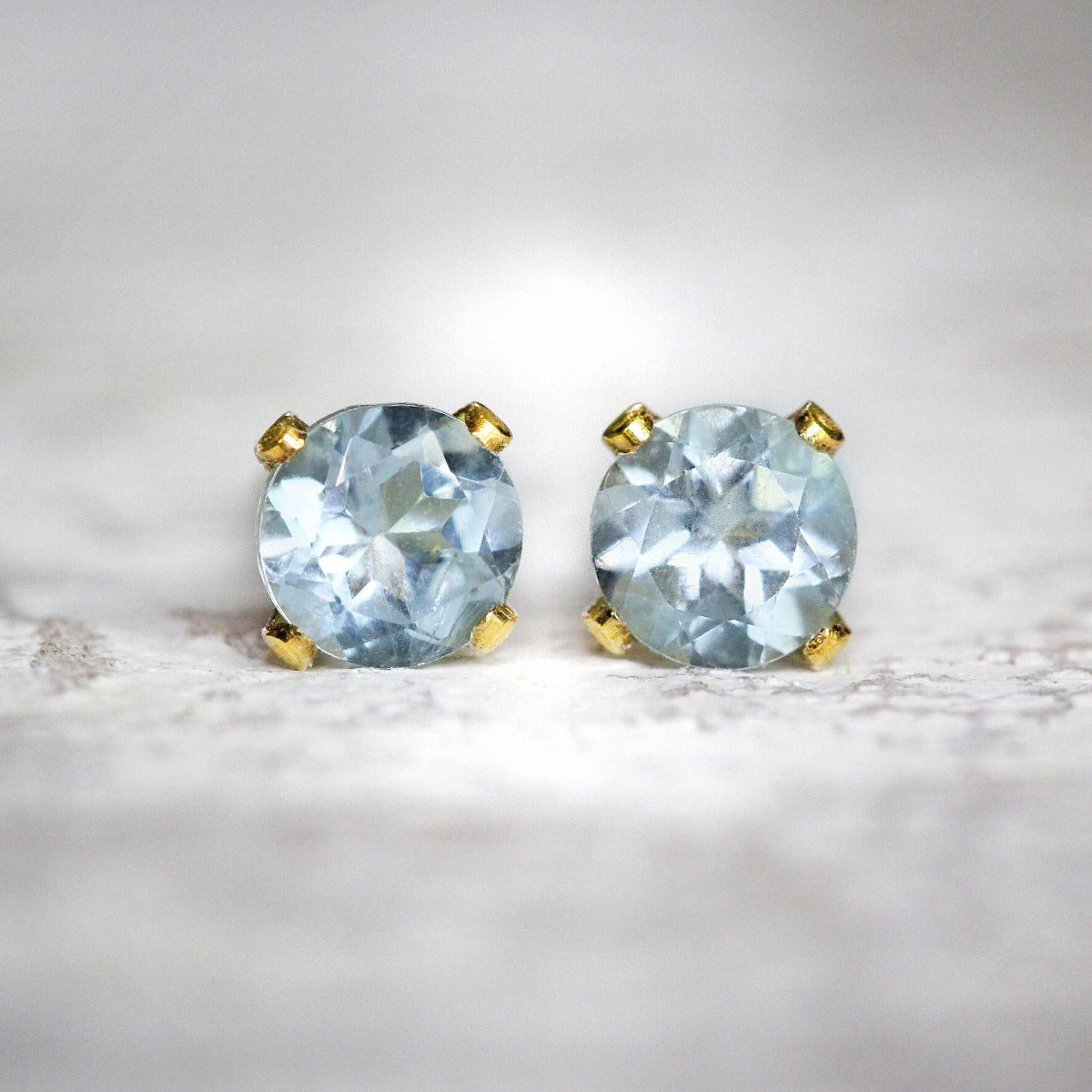 Handmade gold fill stud earrings featuring faceted, pale blue aquamarine gemstones in a prong setting. They are round in shape, with ear post fitting and matching butterfly backs. The earrings are simple & minimal in style, and come in various sizes.