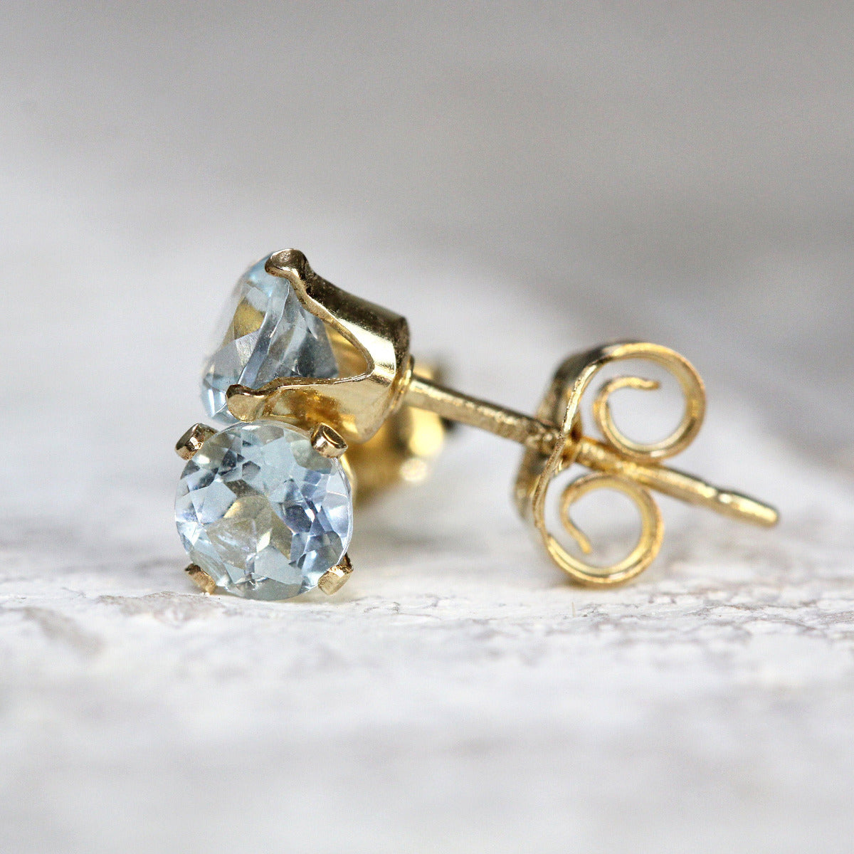 One pair of light blue aquamarine stud earrings. The stone is round in shape with a facted brilliant round cut. The 14k gold fill ear post setting has four prongs holding the gemstone in place, and butterfly push backs to secure the stud on the ear.