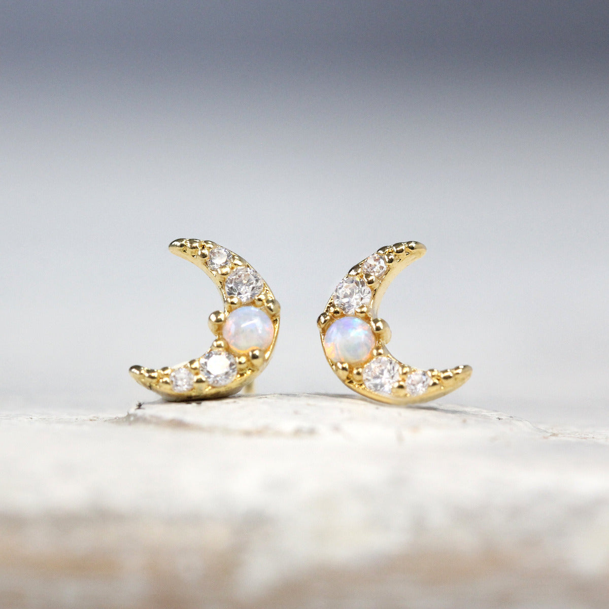 A pair of Crescent Moon Stud earrings with opal and crystal stones set in gold shown on a soft grey background