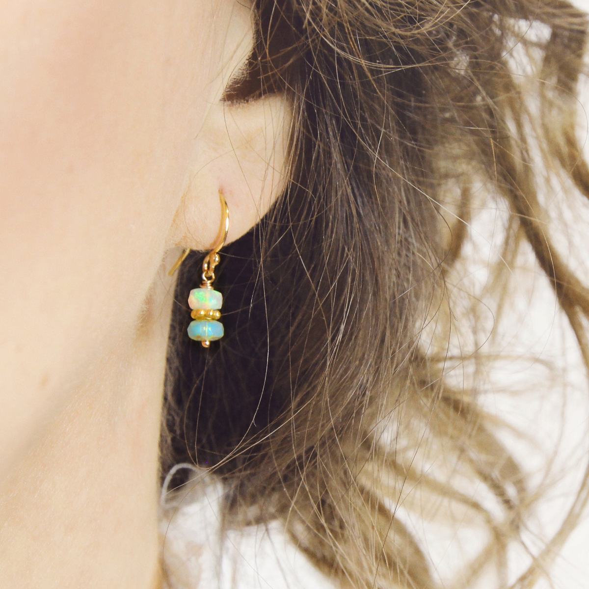 Close-up of a person wearing dainty gold drop earrings with opal stones.