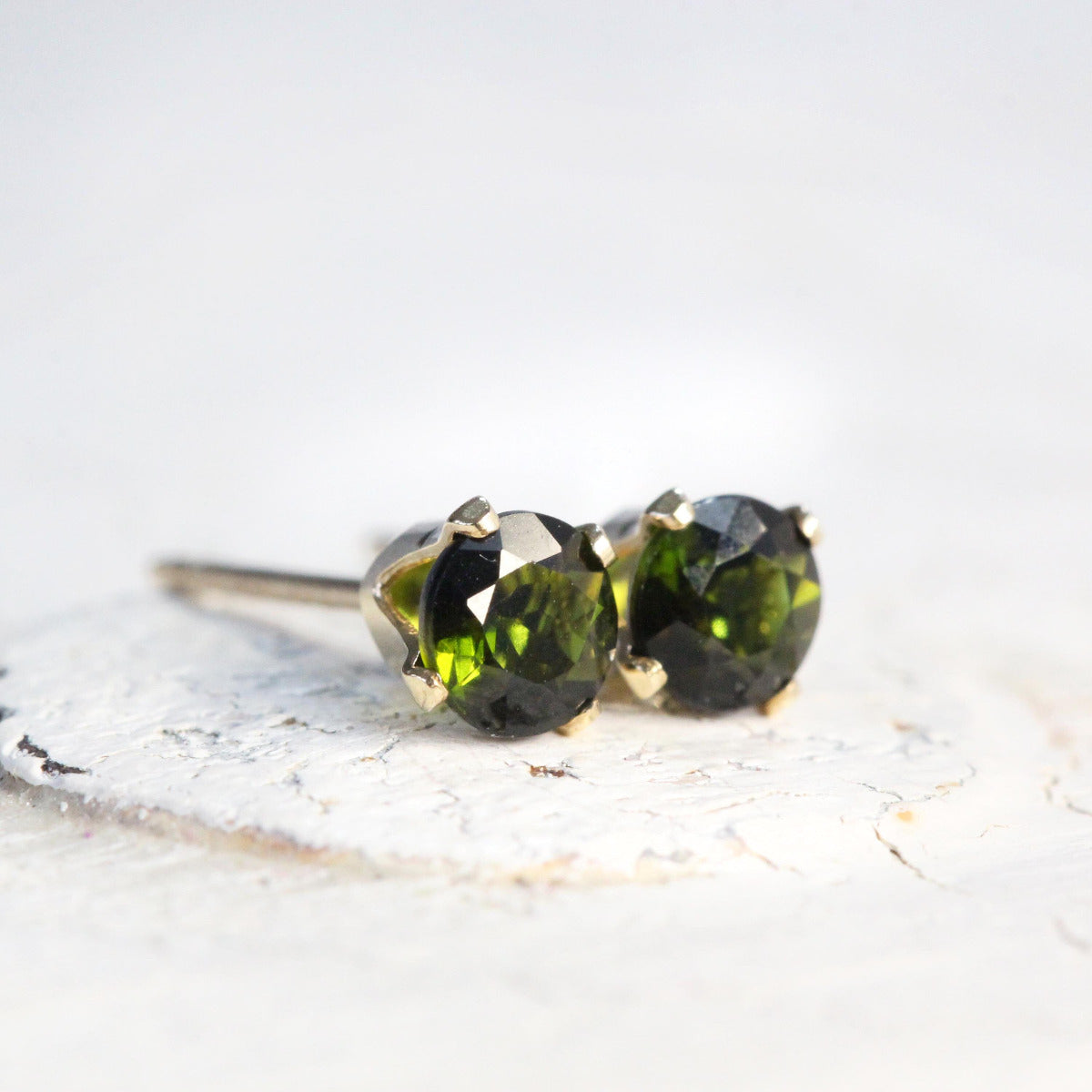 Handmade stud earrings featuring faceted dark olive green tourmaline gemstones set into a silver or gold prong setting. The earrings are round in shape, with ear post fitting and butterfly backs. The earrings are simple and minimal in style.