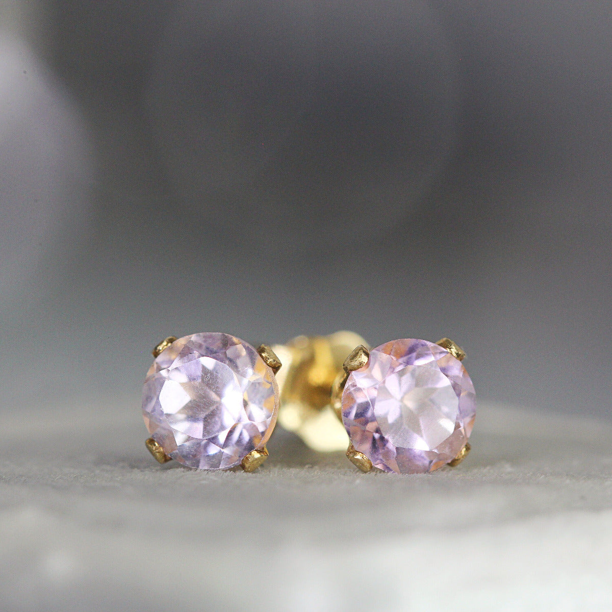 A pair of matching gemstone stud earrings in gold settings. The amethyst gemstone is round, faceted, sparkling and pale lavender purple in colour. The stones are set into 14k gold fill ear posts with a 4 prong setting with matching butterfly backs.