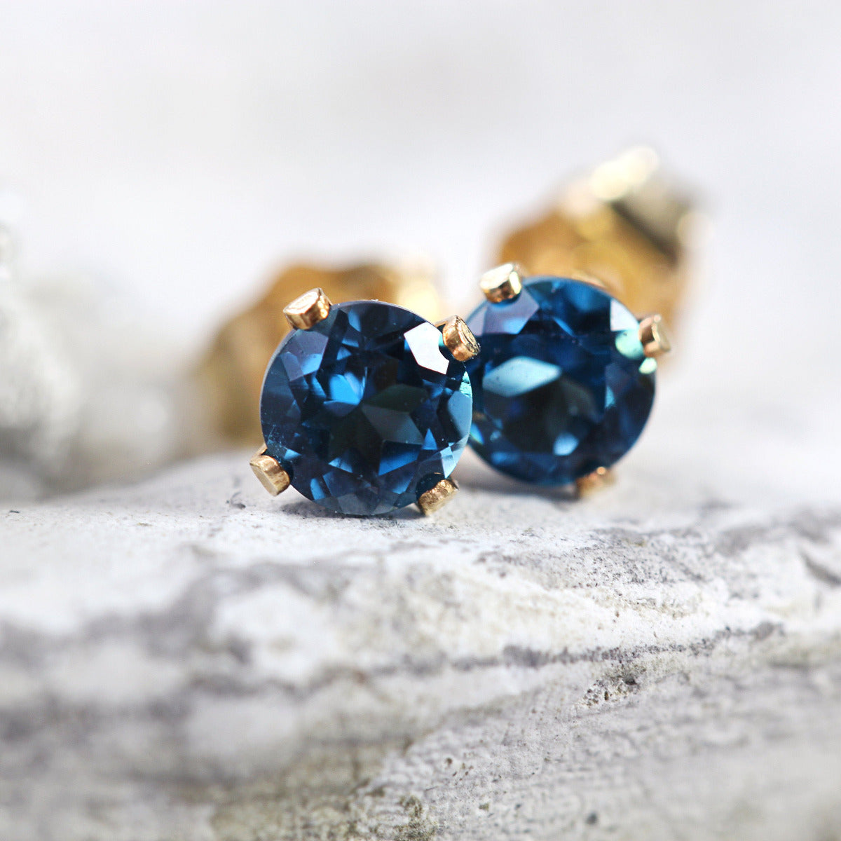 A pair of sparkling stud earrings featuring faceted, sparkling London Blue Topaz gemstones set into a gold prong setting. The earrings are round in shape, with ear post fitting and butterfly backs. The topaz earrings are simple and minimal in style.