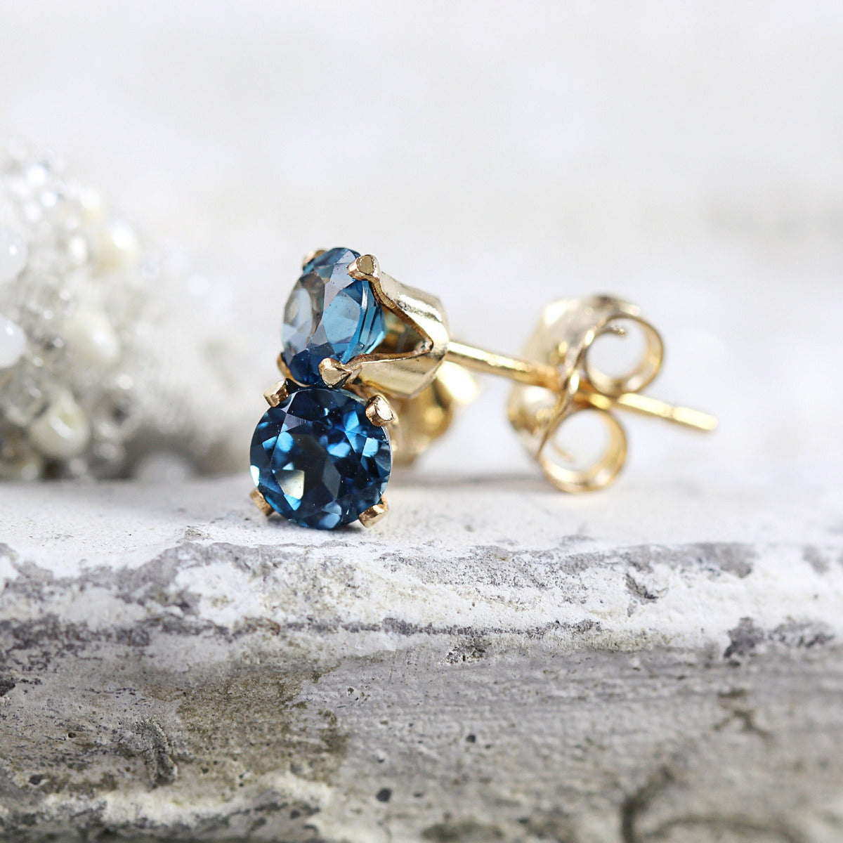 A pair of London Blue topaz stud earrings. The topaz stone is round in shape with a facted cut. The 14k gold fill ear post setting has four prongs holding the gemstone in place, and butterfly push backs to hold the stud securely on the ear.