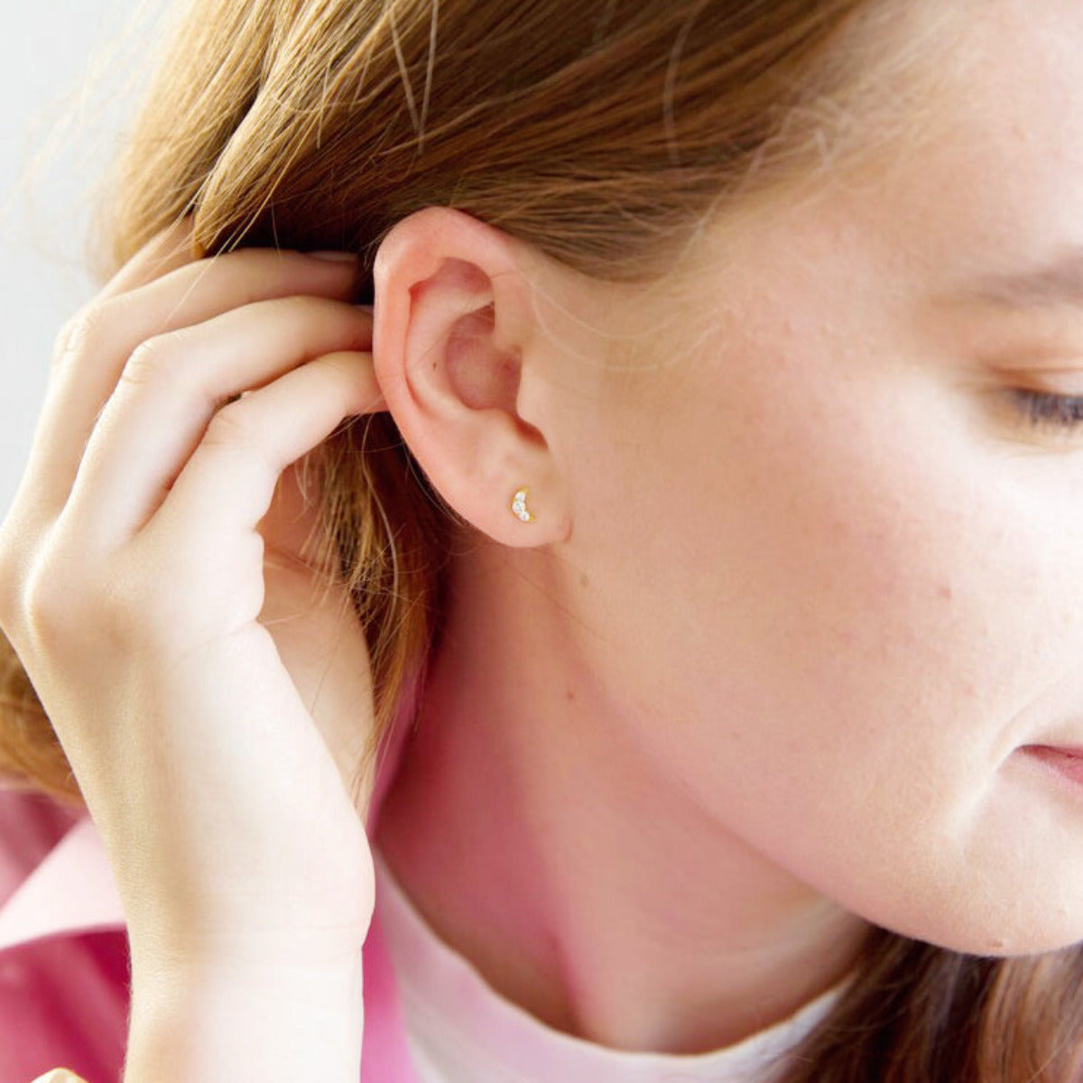 A woman wears a pair of tiny gold crystal moon shaped stud earrings, dressed in magenta pink, holding hair behind ear.