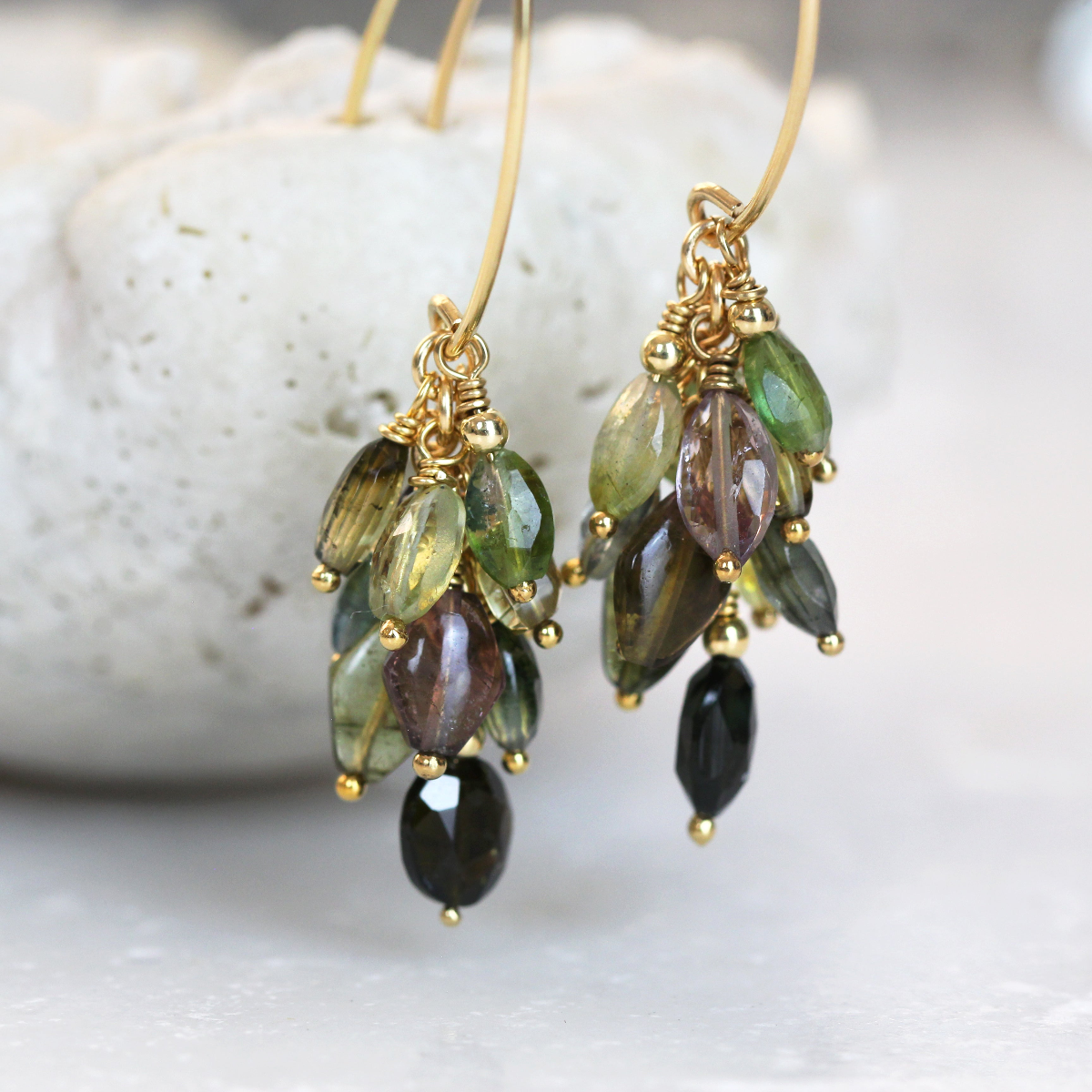 Multi Coloured Tourmaline Cluster Earrings with green, brown, plum and black gemstone beads dangling in a cluster design. The gemstone beads hang from gold ear hooks on a shell bowl against on a light background.