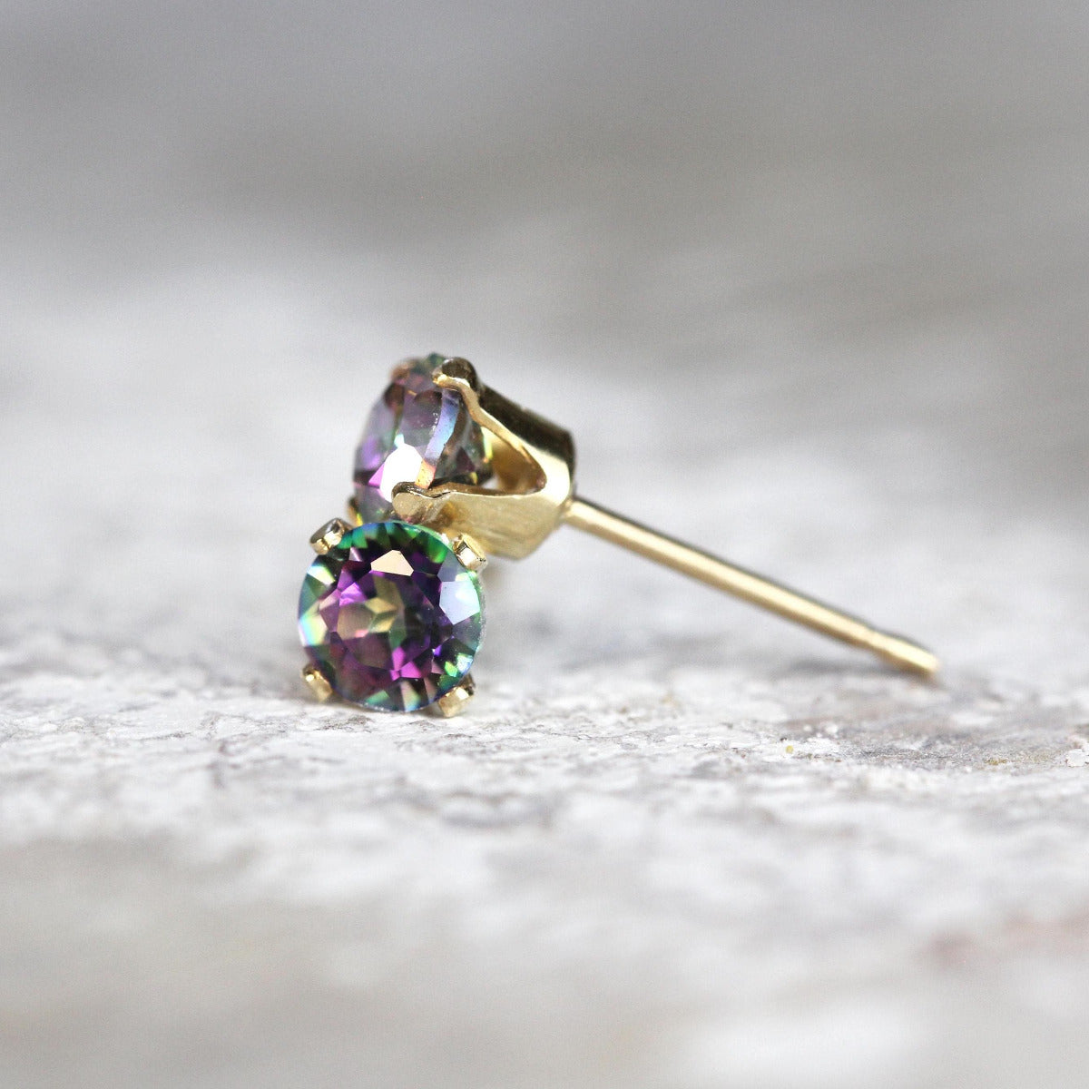 A pair of colourful mystic topaz stud earrings. The topaz stone is round in shape with a facted cut. The 14k gold fill ear post setting has four prongs holding the gemstone in place, and butterfly push backs to hold the stud securely on the ear.