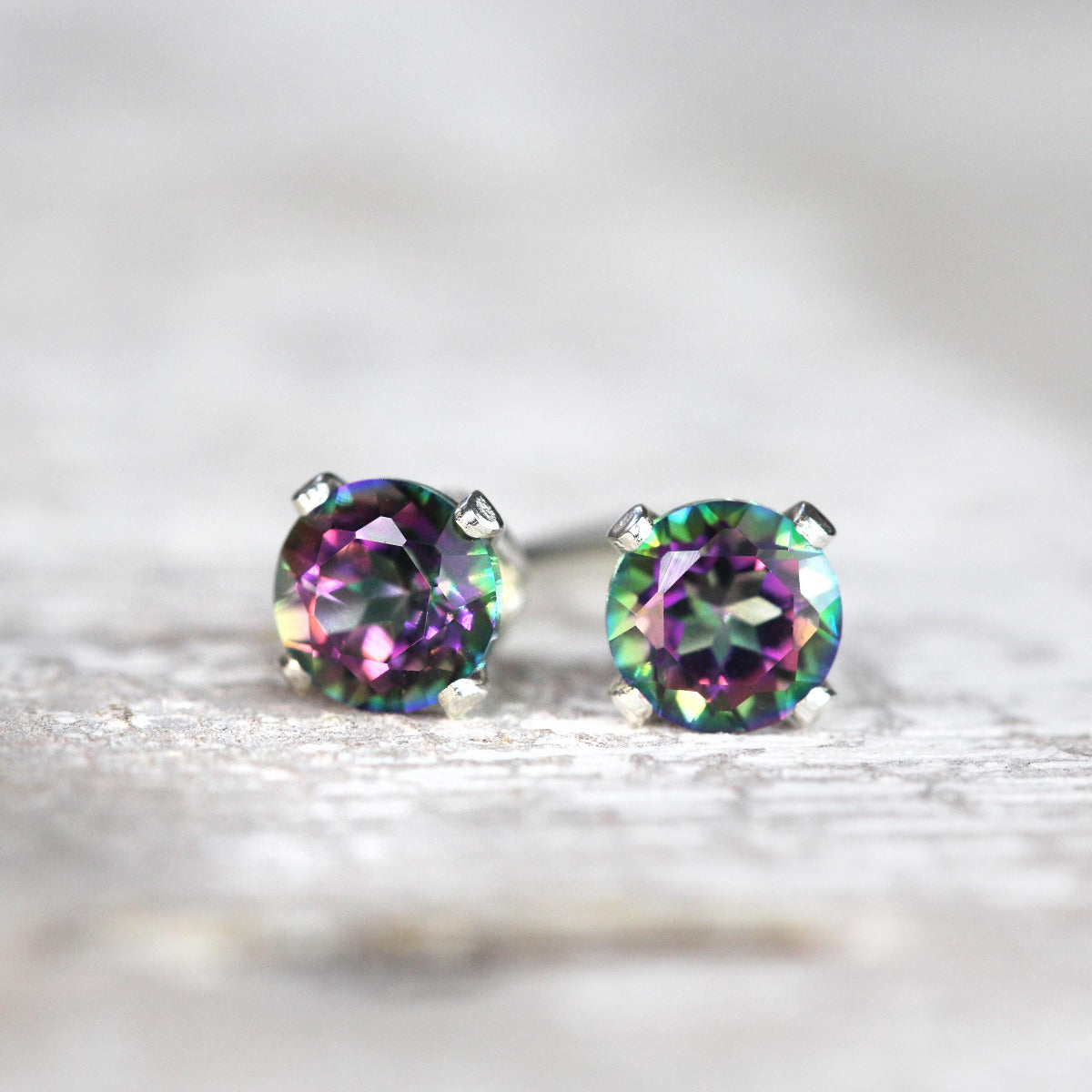A pair of simple minimal stud earrings featuring faceted, sparkling, mystic topaz gemstones. The topaz stones have a dazzling kaleidoscopic colour effect in green, blue, pink and purple, and are set into a sterling silver ear post fitting.