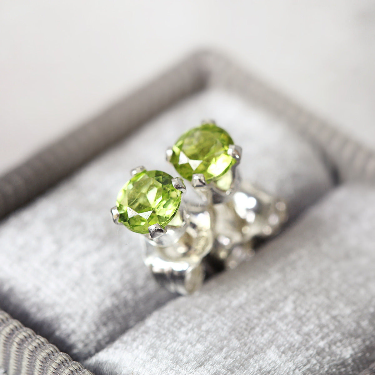 A pair of bright green gemstone stud earrings. The peridot gemstones are sparkling and catch the light. The stones are a brilliant round cut with lots of facets, and set into sterling silver four prong settings. Simple, minimal, round stud earrings.