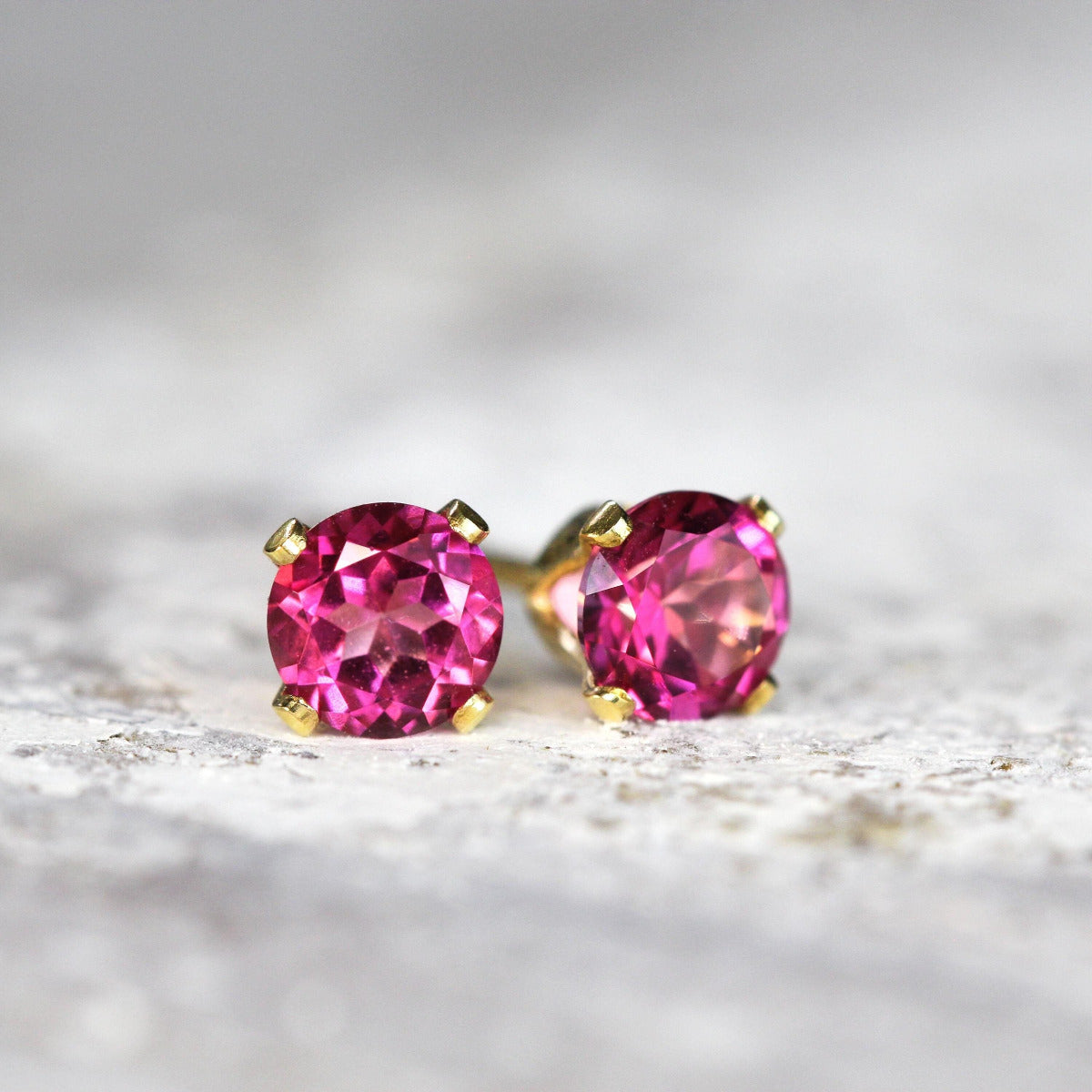 Gold stud earrings featuring faceted, sparkling, hot bright pink topaz gemstones set into a gold fill prong setting. The earrings are a brilliant round cut, with ear post fitting and butterfly backs. The earrings are simple and minimal in style.