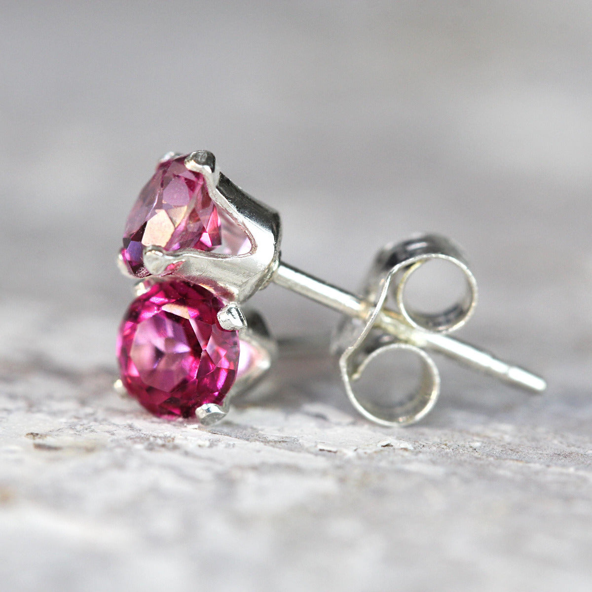 A pair of mystic pink topaz stud earrings. The magenta topaz stone is round in shape with a facted cut. The 14k gold fill ear post setting has four prongs holding the gemstone in place, and butterfly push backs to hold the stud securely on the ear.