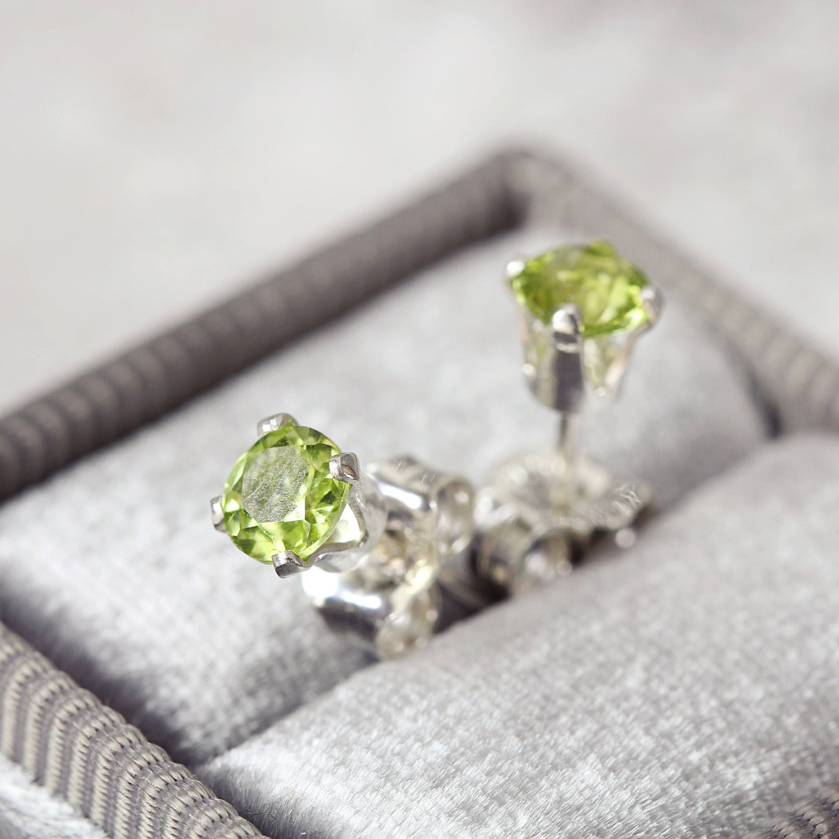 A pair of bright green gemstone stud earrings. The peridot gemstones are sparkling and catch the light. The stones are a brilliant round cut with lots of facets, and set into sterling silver four prong settings. Simple, minimal, round stud earrings.