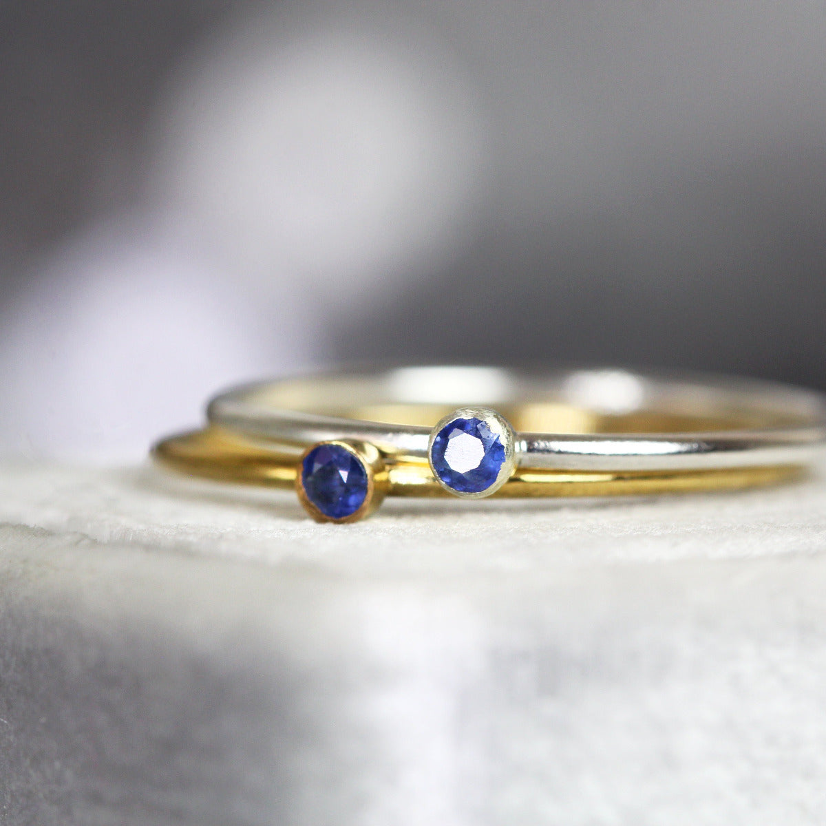 Two stacking rings with blue sapphire gemstones on a textured surface. One gold ring and one silver ring.