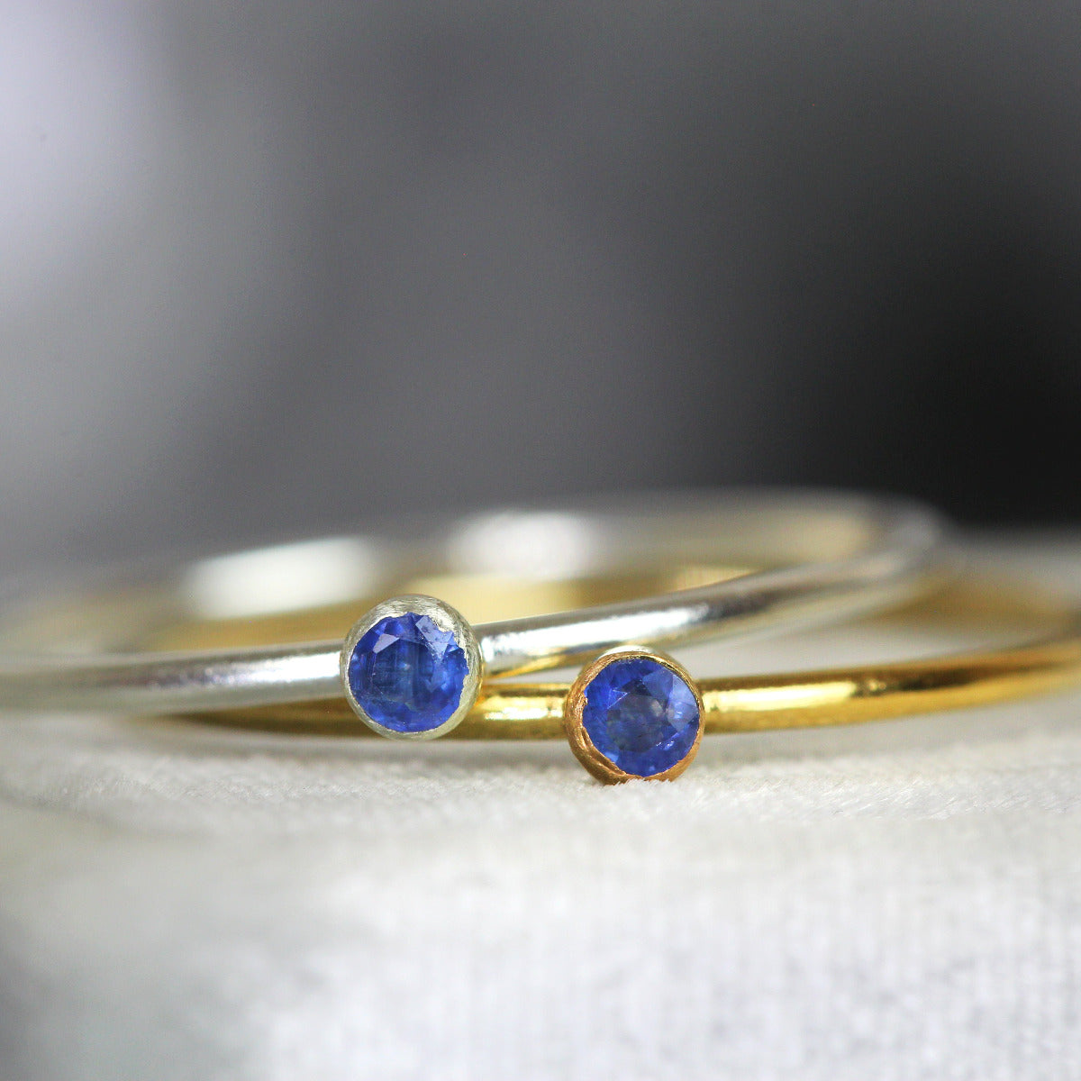 Two rings, one gold and one silver, each with a blue sapphire gemstone on a neutral background.