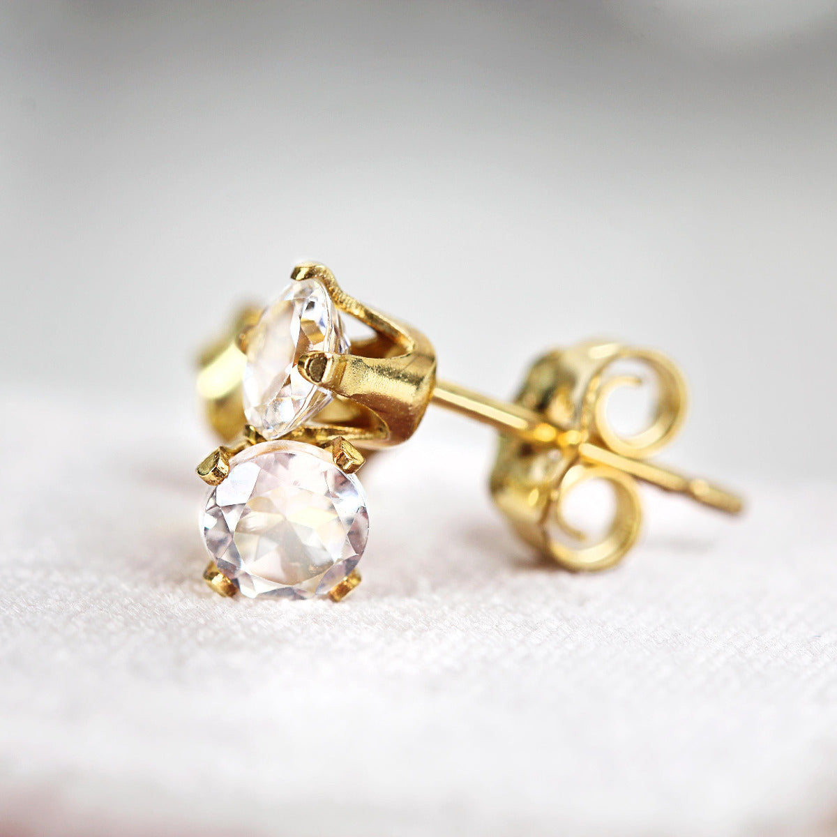 A pair of white topaz stud earrings. The topaz stones are round in shape with a facted cut. The 14k gold fill ear post setting has four prongs holding the gemstone in place, with butterfly push backs to hold the stud securely on the ear.