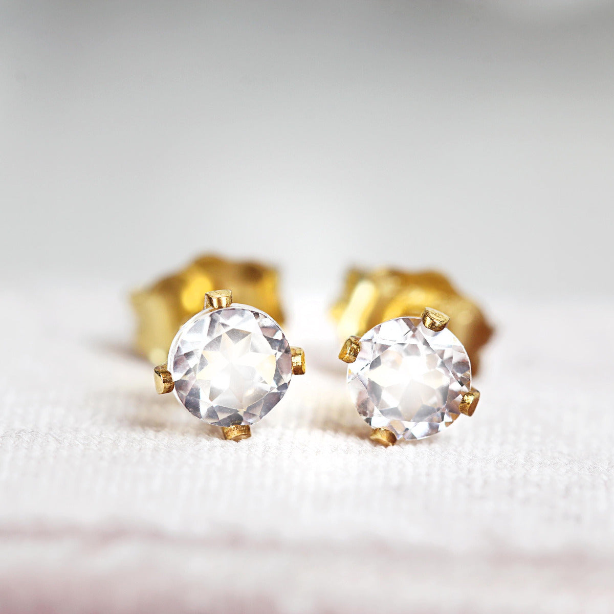 A pair of stud earrings featuring faceted, sparkling White Topaz gemstones set into a gold prong setting. The earrings are round in shape, with ear post fitting and butterfly backs. The topaz earrings are simple and minimal in style.