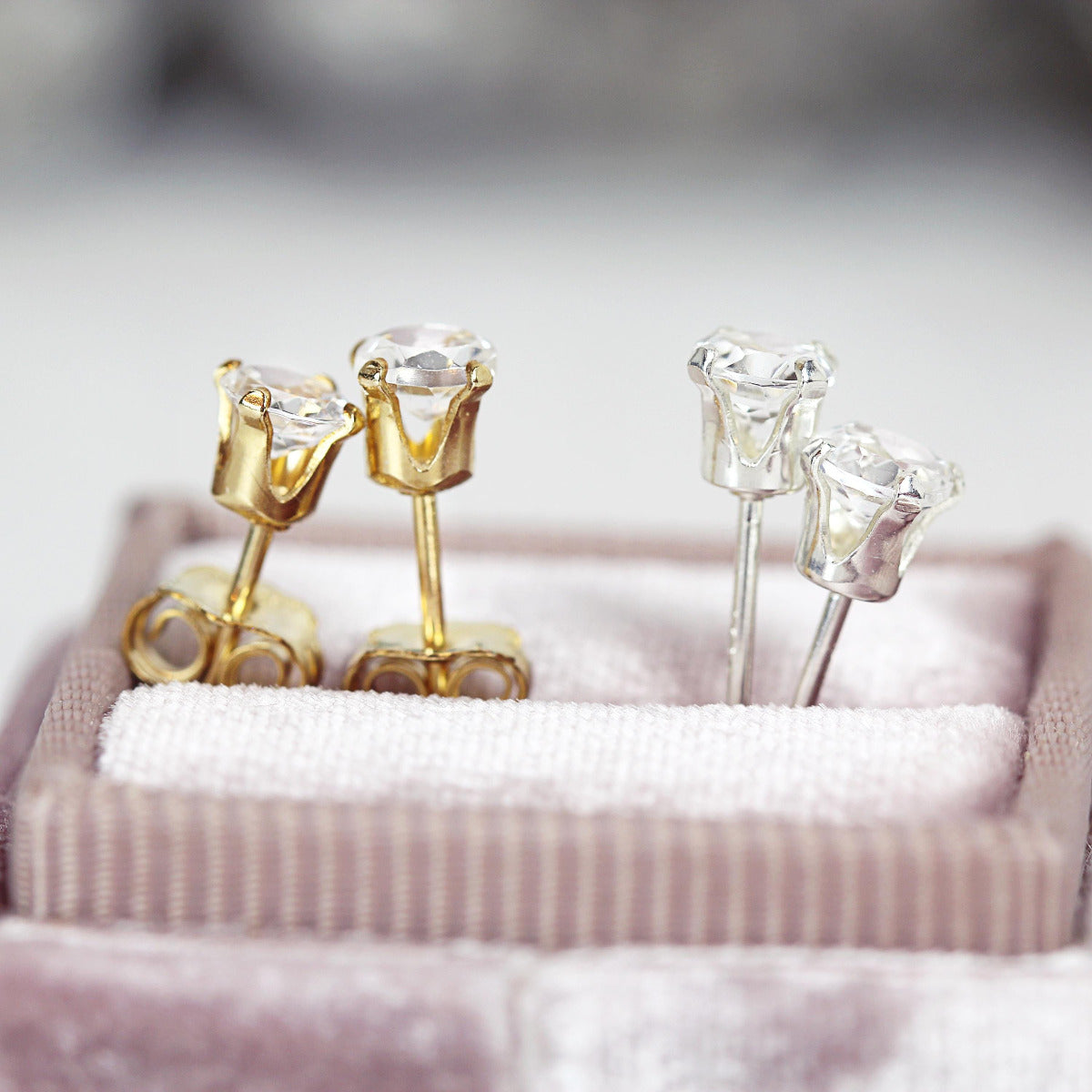 Two pairs of stud earrings shown side by side. They feature sparkling white topaz gemstones in a  brilliant round cut. One pair is set into 14k gold fill ear posts and the other pair is set in sterling silver. The studs are round, simple and minimal.