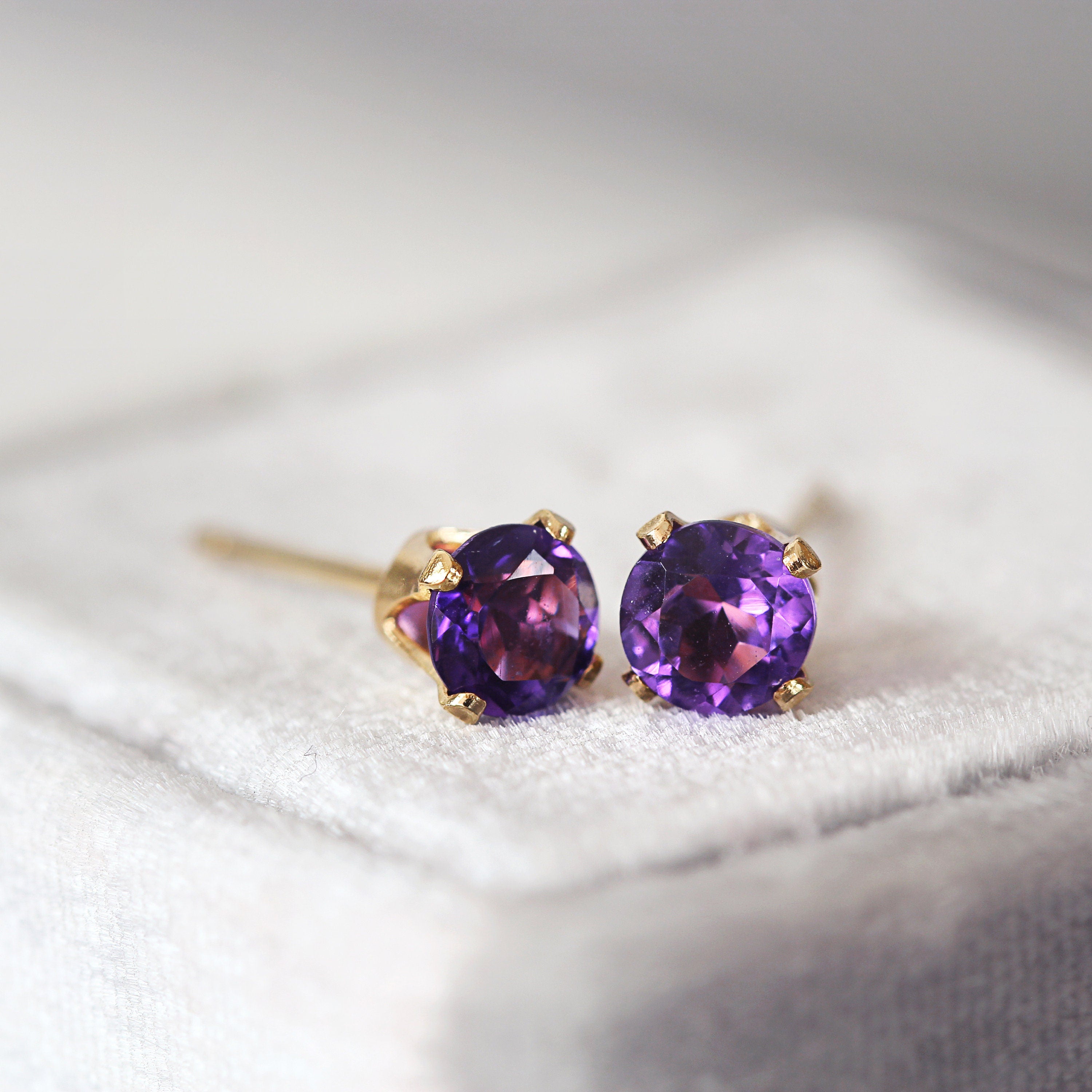 A pair of deep purple amethyst stud earrings. The stone is round in shape with a facted brilliant round cut. The 14k gold fill ear post setting has four prongs holding the gemstone in place. Simple, minimal everyday or evening amethyst earrings.