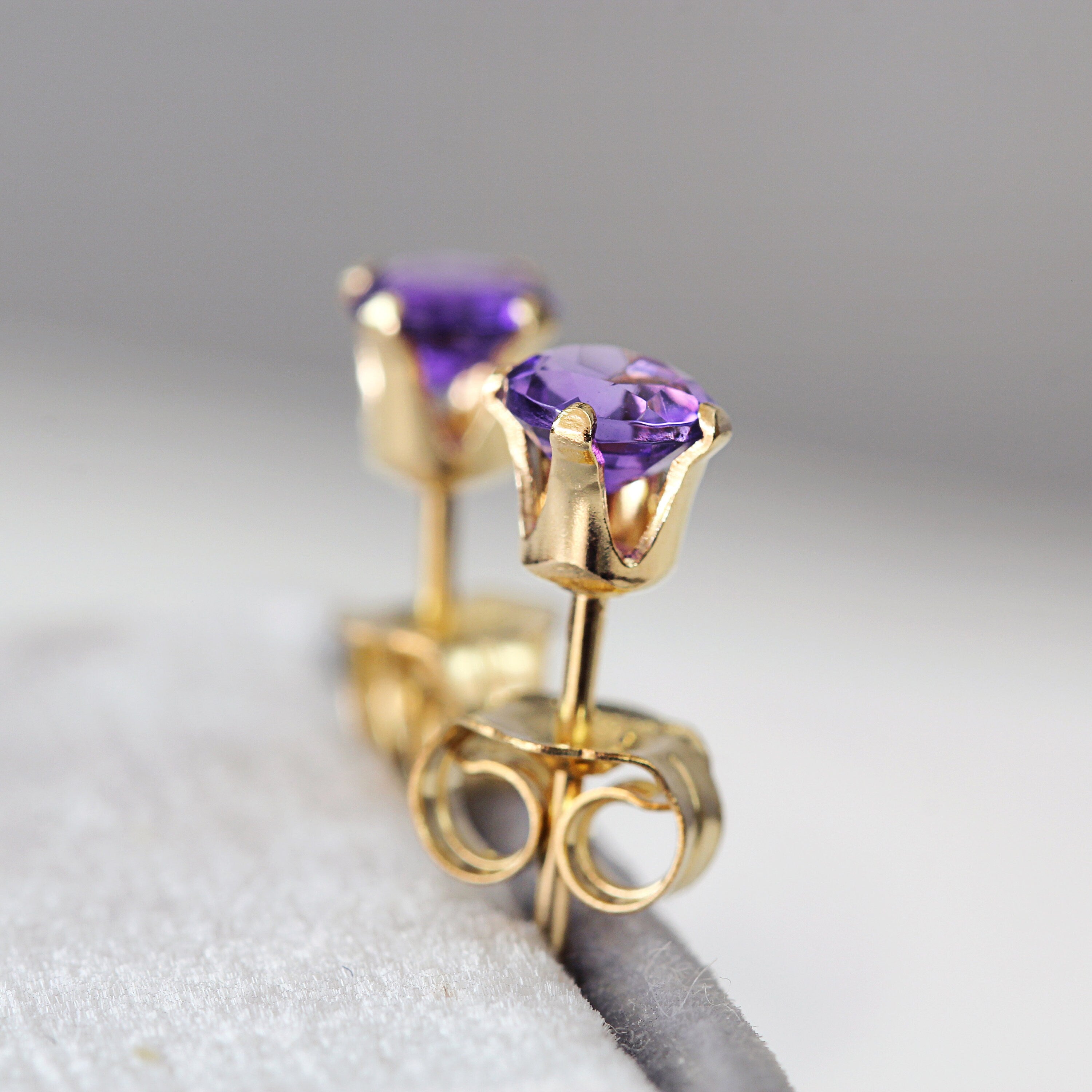 A pair of deep purple amethyst stud earrings. The stone is round in shape with a facted brilliant round cut. The 14k gold fill ear post setting has four prongs holding the gemstone in place, and butterfly push backs to secure the stud on the ear.